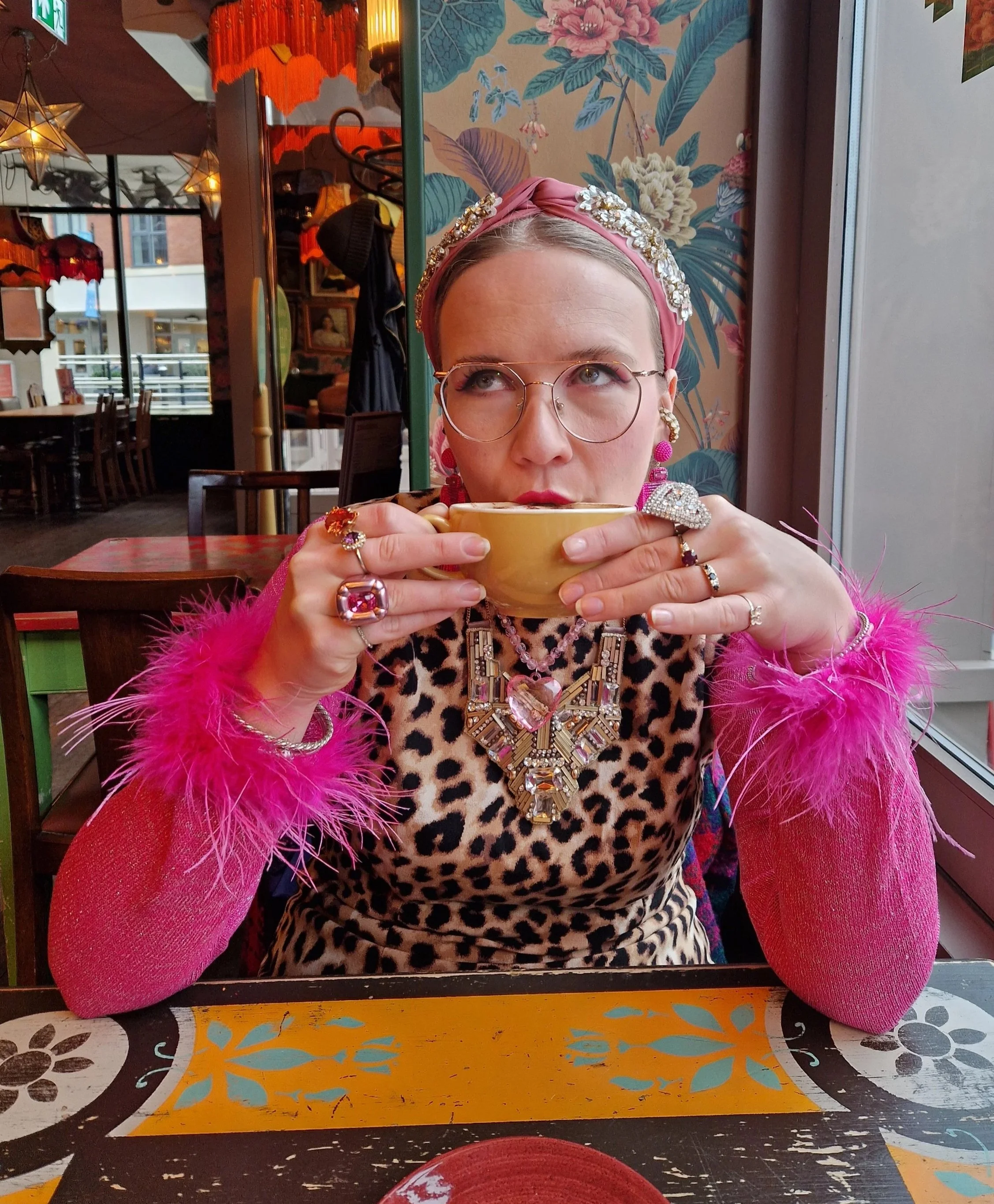 A woman with platinum hair wearing pink headband, glasses, and colorful jewelry in a floral-patterned room, holding a yellow bowl close to her lips.