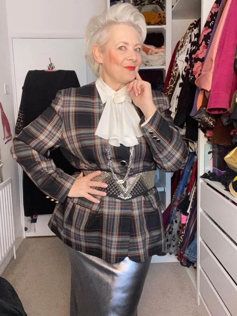 A woman with short, white hair posing confidently in a walk-in closet filled with clothes and accessories, wearing a plaid blazer, a metallic skirt, and statement jewelry.