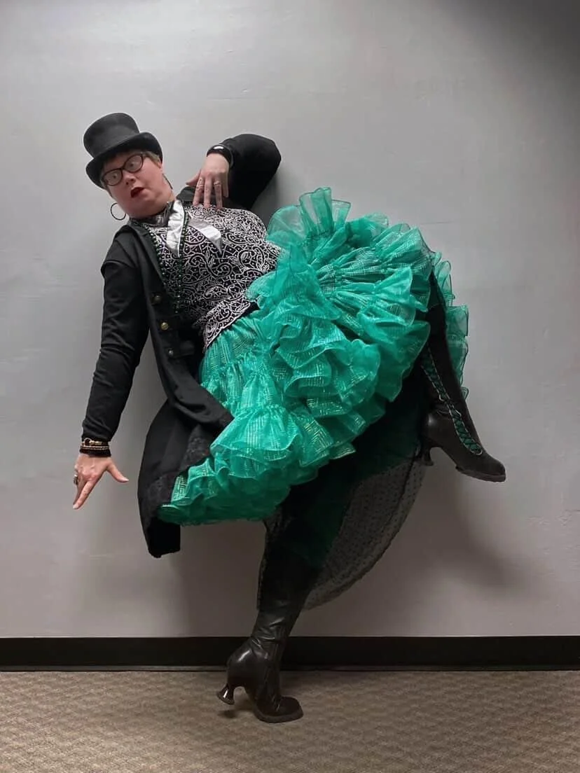 Person dressed in a black blazer, leopard print top, teal ruffled skirt, and tall black leather boots, posing against a gray wall with a playful attitude, wearing glasses, earrings, and a top hat.