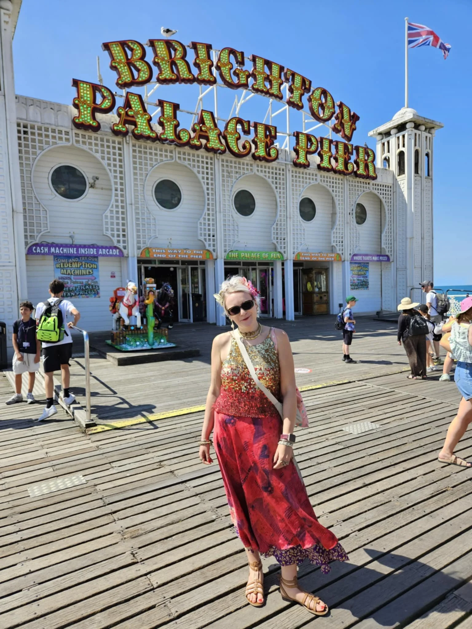 A woman wearing a sleeveless dress with a red and purple pattern, standing on a wooden pier in front of an amusement park entrance, smiling at the camera. The amusement park sign reads 'Brighton Palace Pier,' with banners and people in the background.