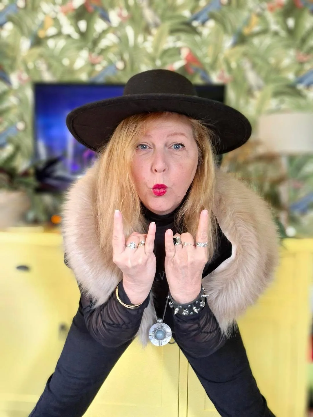A woman with blonde hair wearing a large black hat, a fur stole, and multiple rings and bracelets, making a rock-and-roll hand gesture with lips pursed.