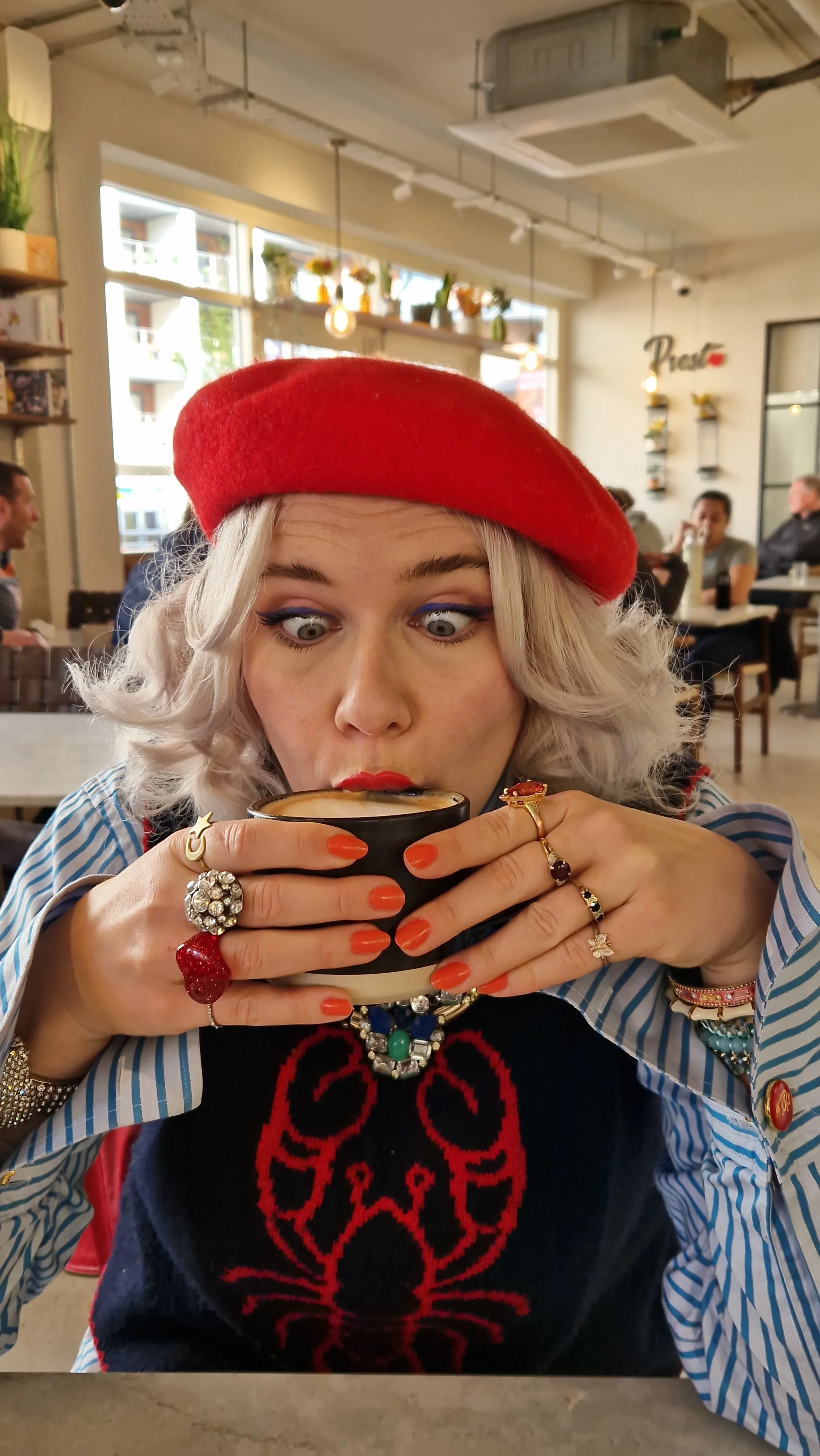 A woman with platinum blonde hair wearing a red beret and colorful jewelry is holding a black and white mug and sipping a hot beverage in a cozy cafe. The cafe has large windows, decorative plants, and several people in the background.