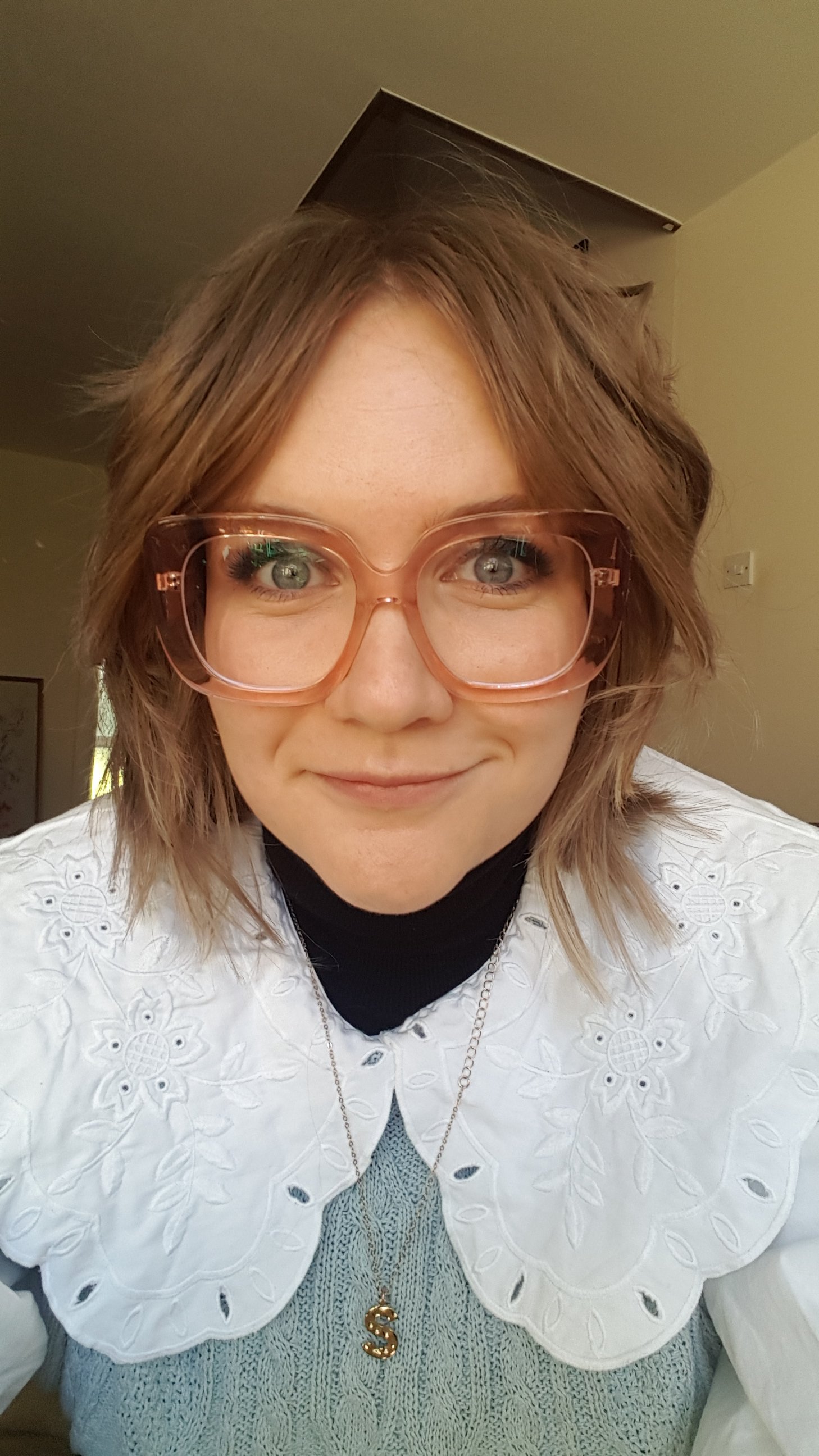 Close-up of a woman with shoulder-length wavy brown hair wearing pink glasses, a white embroidered blouse, a black turtleneck, and a gold dollar sign necklace, smiling at the camera indoors.