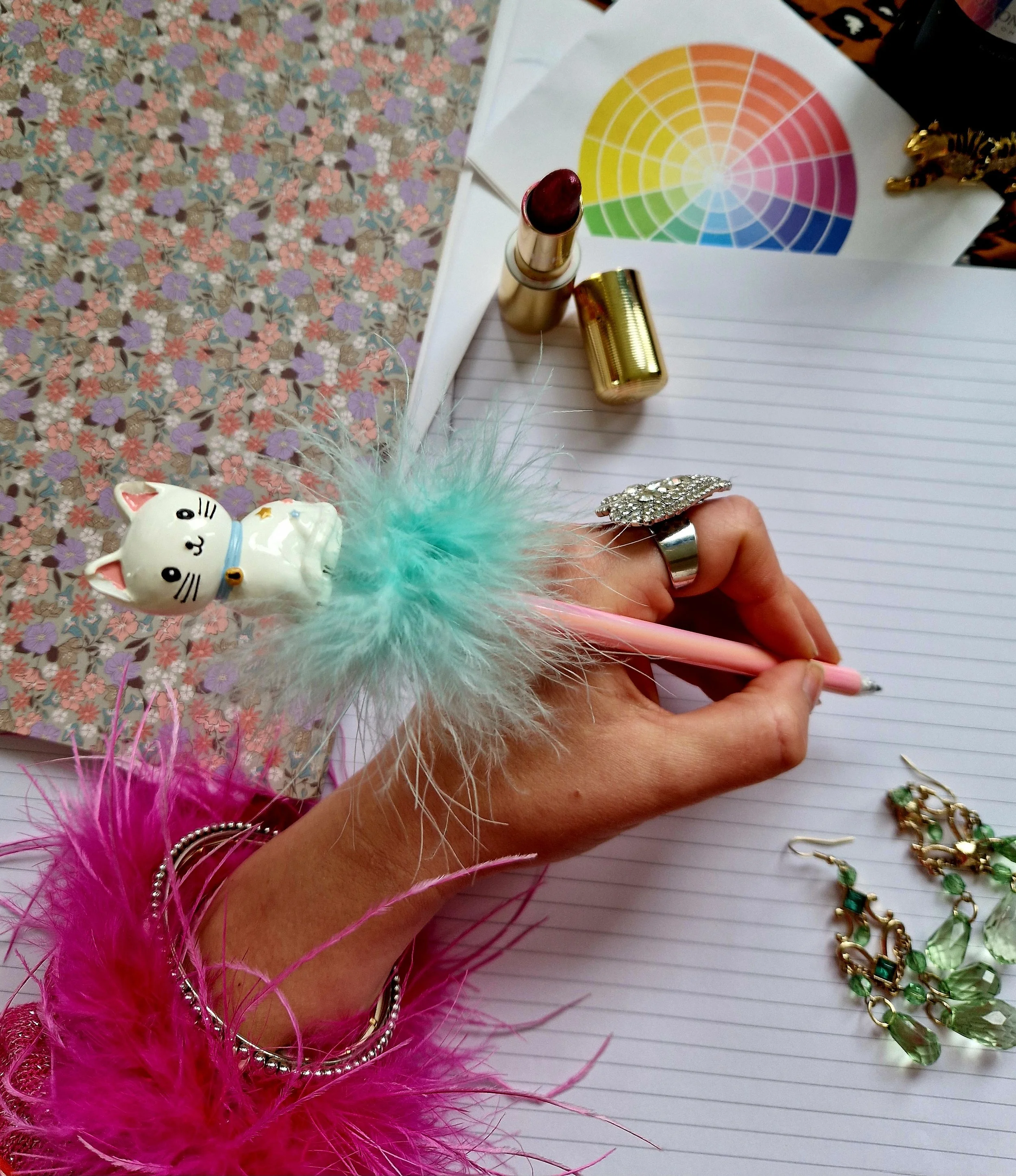 A collection of jewelry items, a notebook with a pink pen, a lipstick, a color wheel, and a ceramic cat figurine with a fluffy pink feather, all placed on a floral patterned surface.