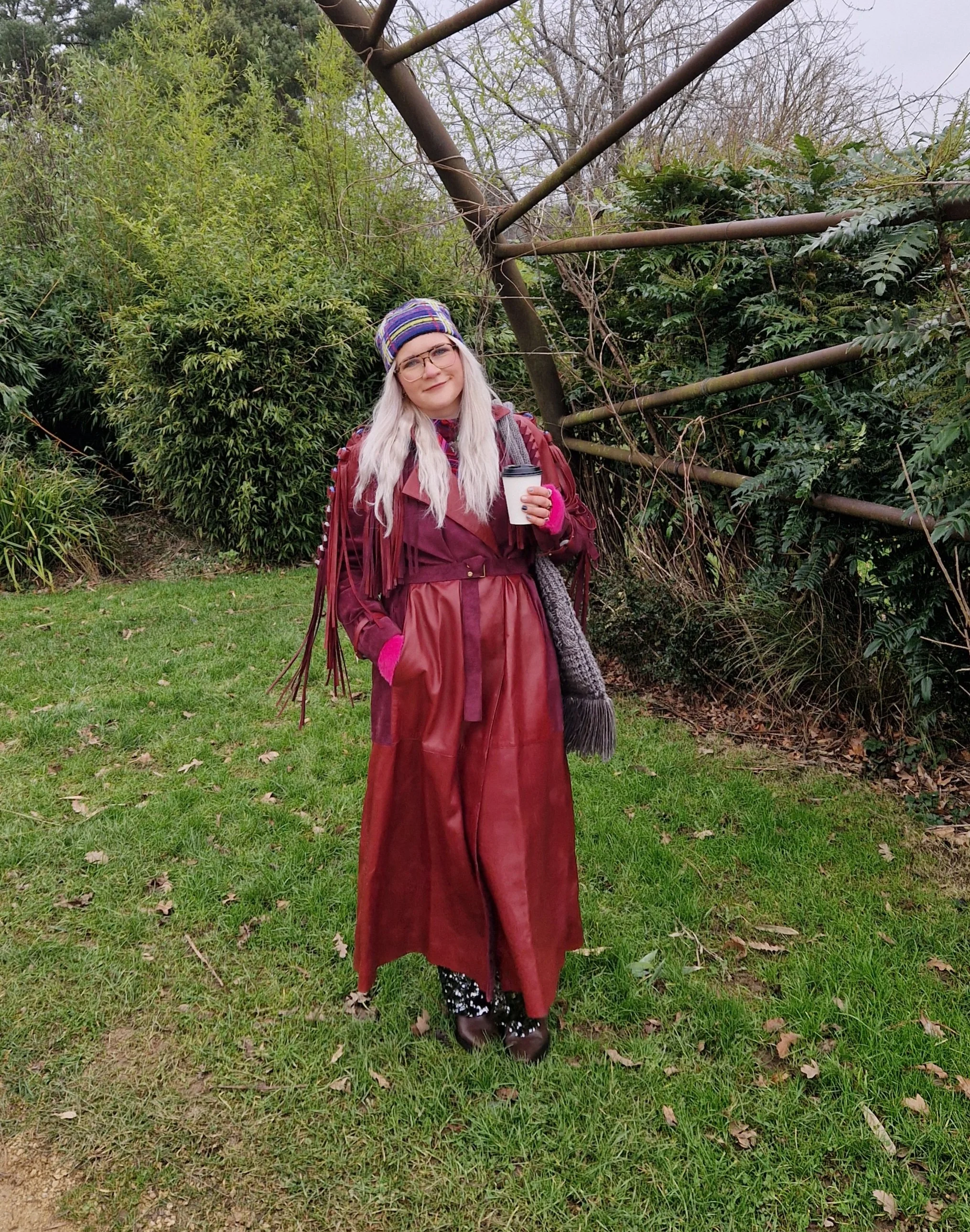 A woman with long, white hair, glasses, and a colorful knit hat holding a white paper coffee cup stands outdoors in a park or garden, surrounded by green plants, bushes, and trees, wearing a long brown trench coat and pink gloves.
