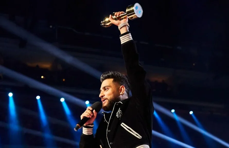A man on stage holding a shiny award trophy high above his head while speaking into a microphone, with blue stage lights shining in the background.