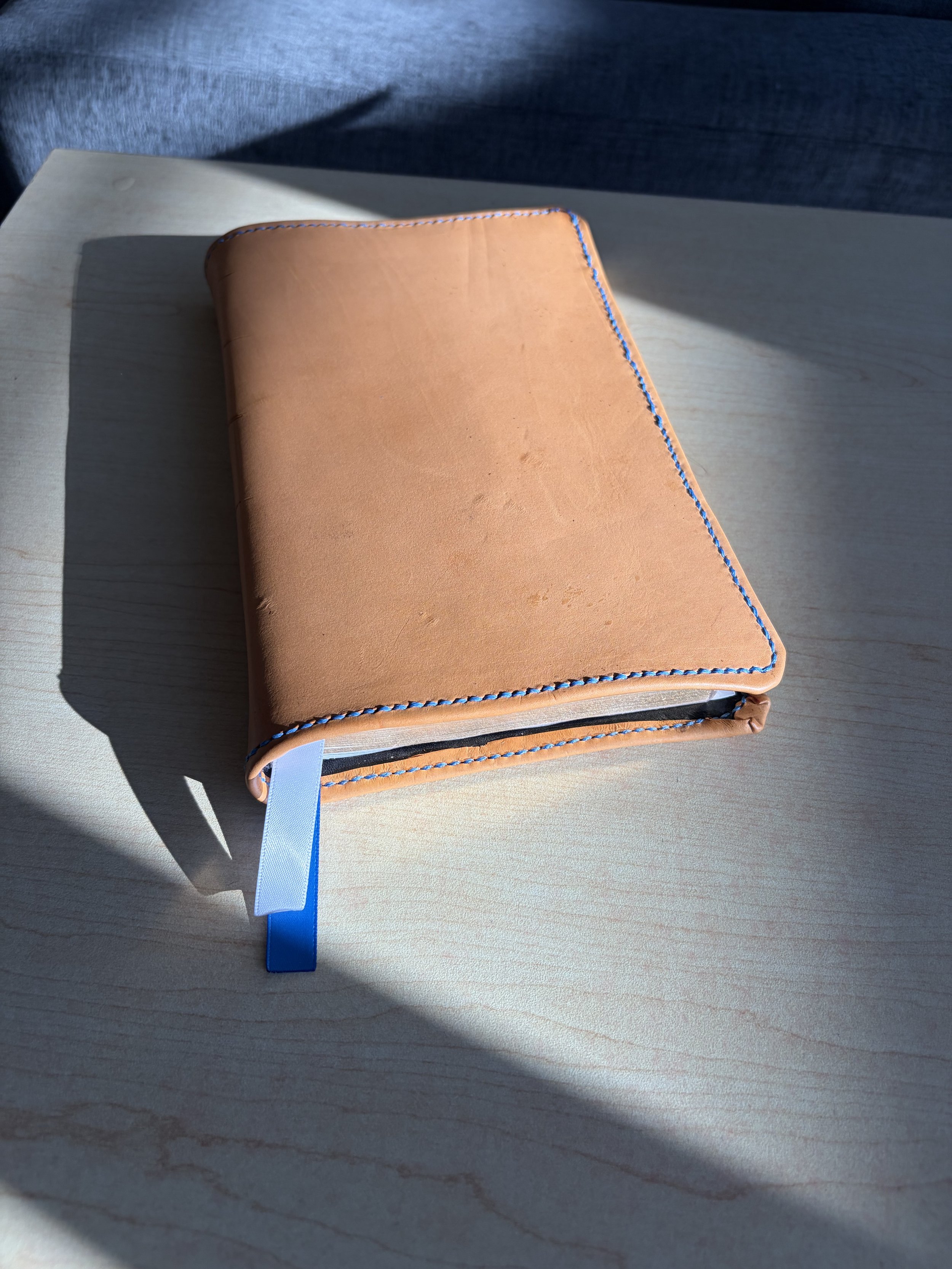 Light brown leather-bound notebook with blue and white ribbon markers on a light-colored wooden surface, partially illuminated by sunlight.