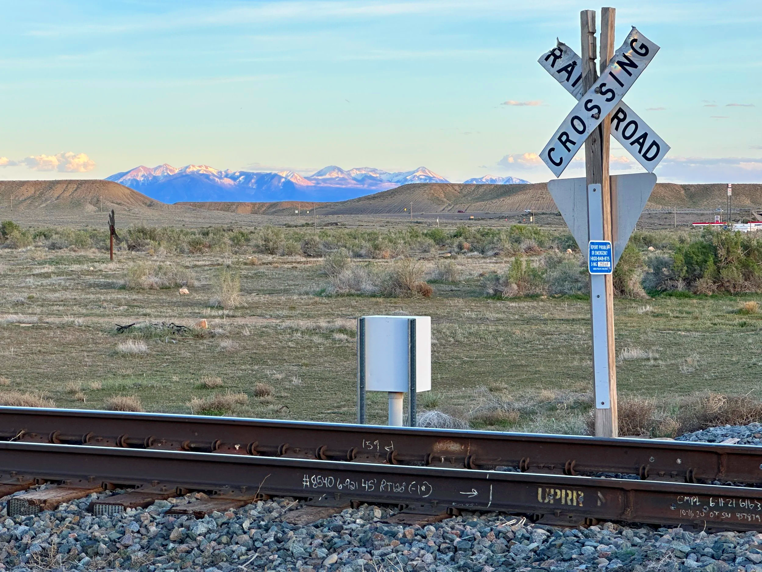 View from a train window showing railroad tracks in the foreground, with a barrier and a mountain range with snow-capped peaks in the background. There is a railroad crossing sign on the right side of the image.