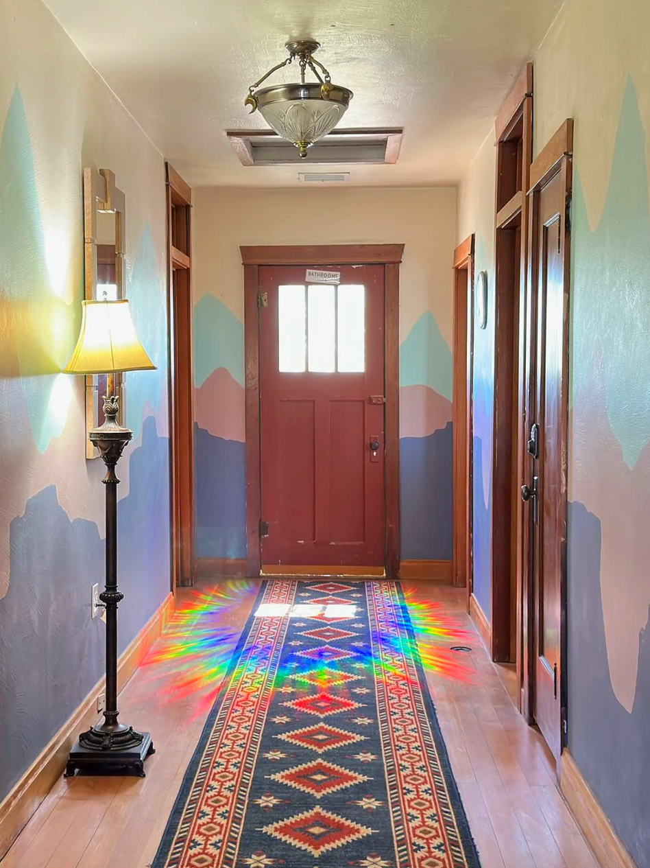 Hallway with wooden trim, colorful mountain mural on walls, patterned runner rug, standing lamp with yellow shade, ceiling light fixture, and door marked 'Bathrooms' at end of hall. Light from outside creates rainbow patterns on the floor.