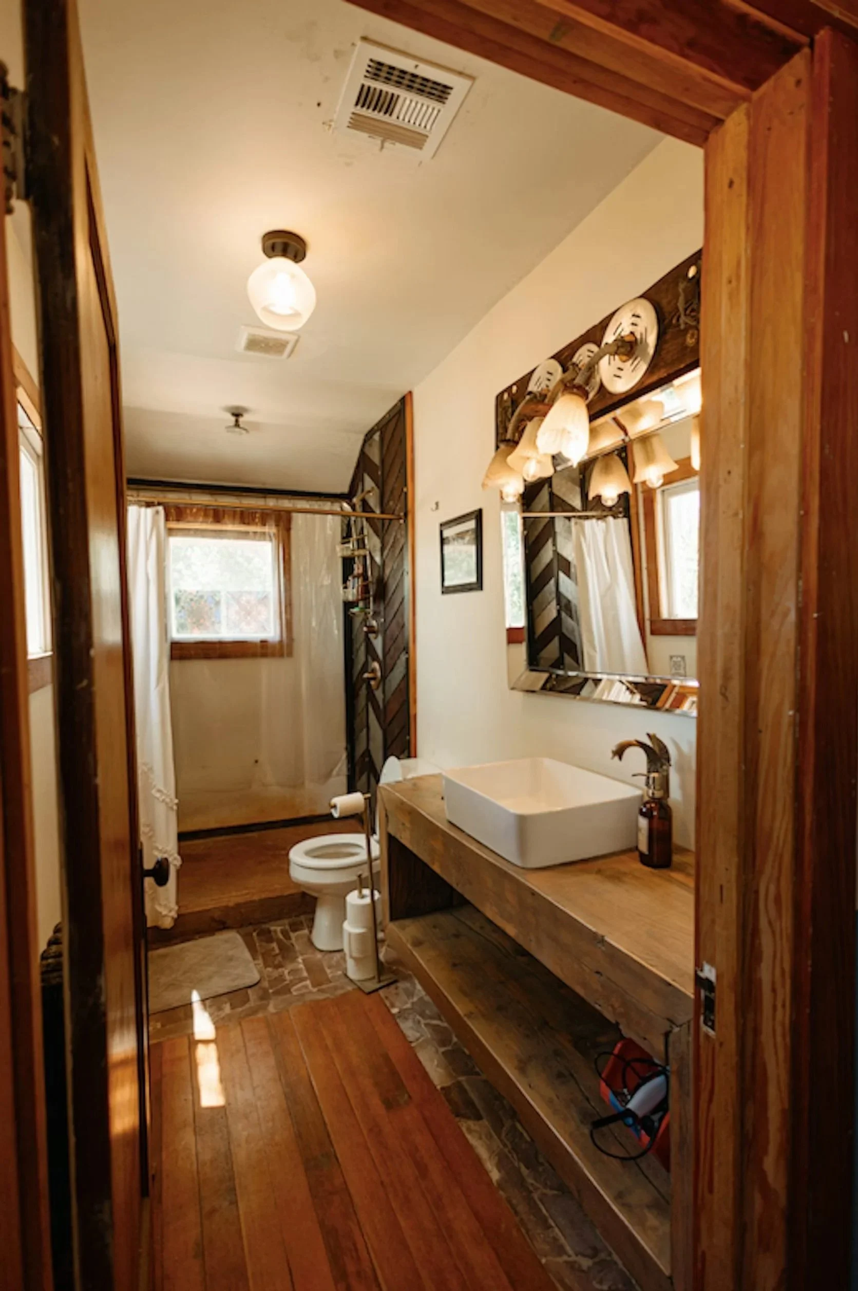 A rustic bathroom with wooden accents, white vessel sink, and a large mirror, featuring a window with white curtains, a toilet, and various lighting fixtures in a cozy, cabin-style setting.