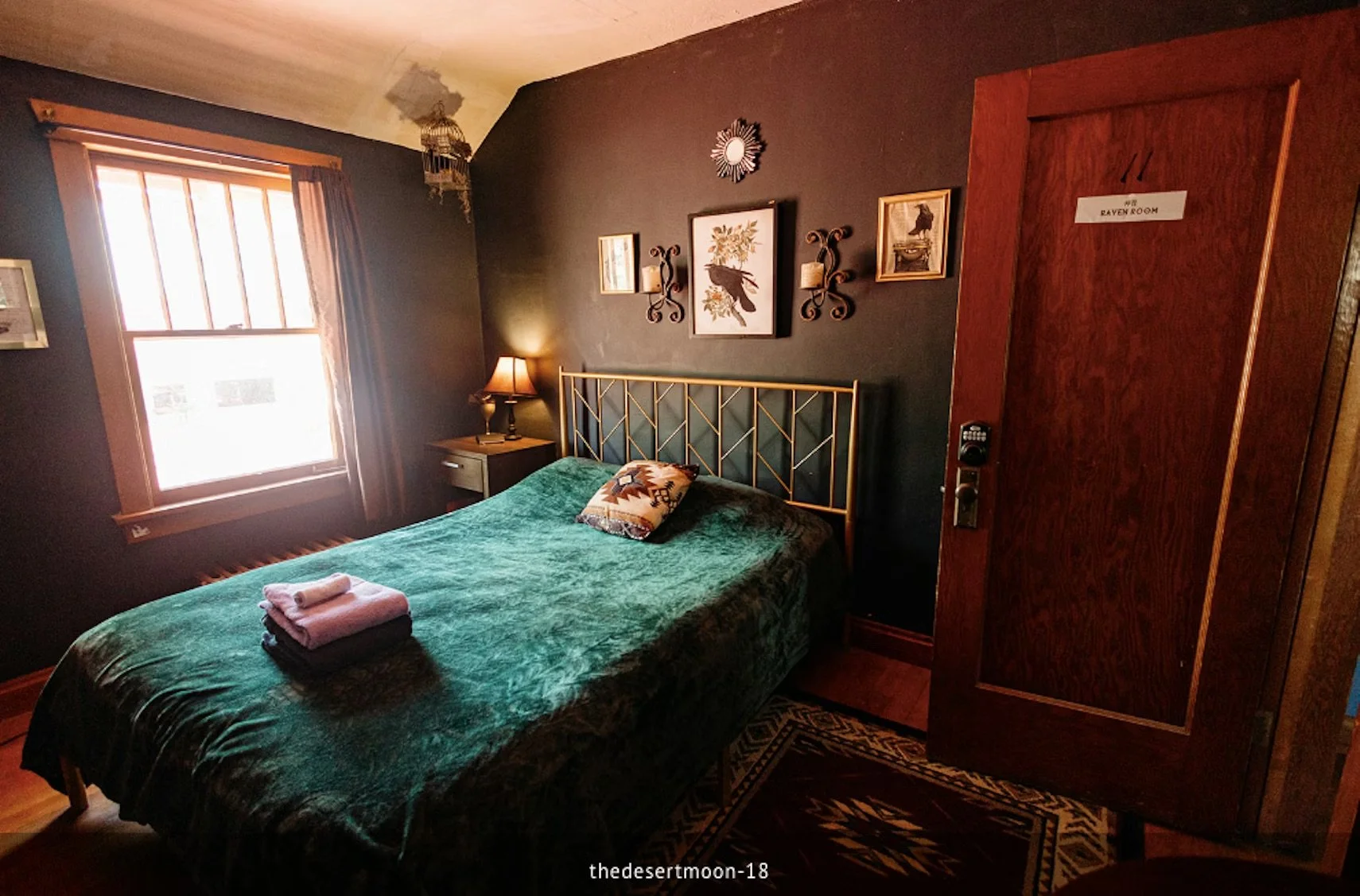 A cozy bedroom with a bed covered in a green blanket and a decorative pillow, a window with wooden blinds and curtains, a small nightstand with a lamp, framed artwork on the dark-colored wall, and a wooden door marked 'The RAVEN ROOM.'