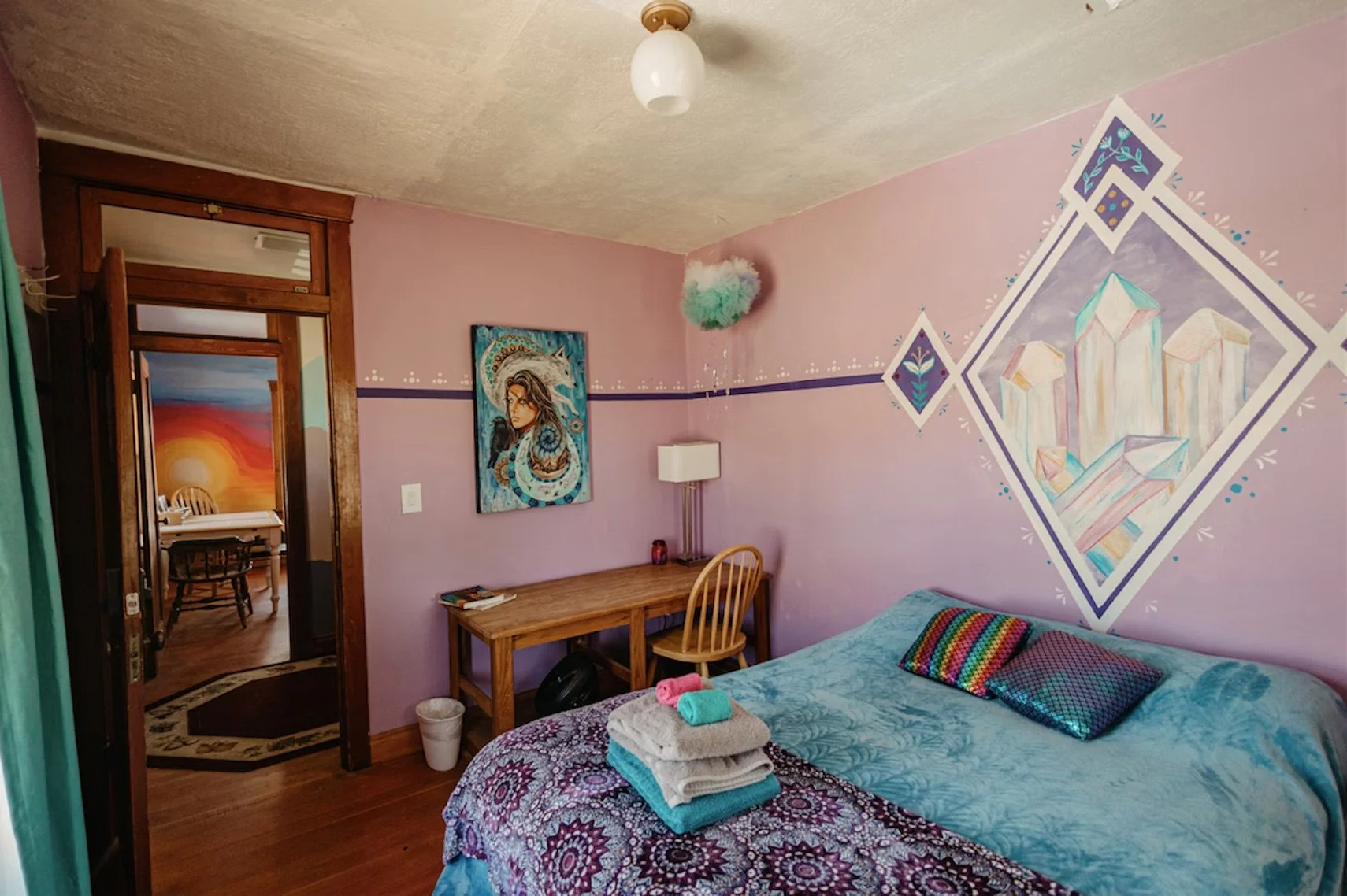 A cozy bedroom with pink walls and colorful artwork, including a mural of a cityscape and a fantasy woman, a wooden desk with a chair, a bed with folded towels on it, and a glimpse into another room with sunset painting.