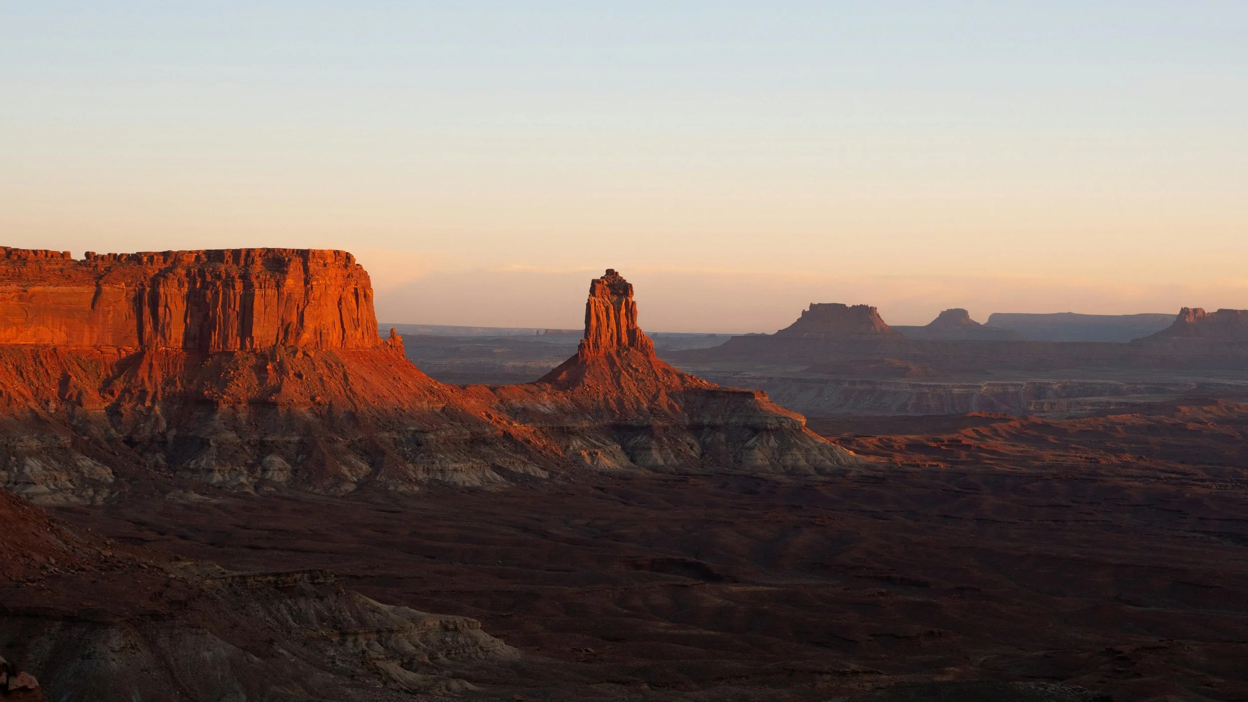 A desert landscape at sunset with red rock formations and mesas in the distance.
