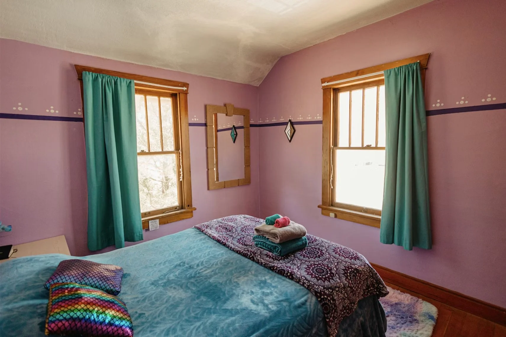A cozy bedroom with pink walls, two windows with wood trim, and turquoise curtains. A bed with a blue bedspread and colorful pillows is in the foreground, with folded towels on it. There is a wall mirror and two diamond-shaped decorations on the wall.