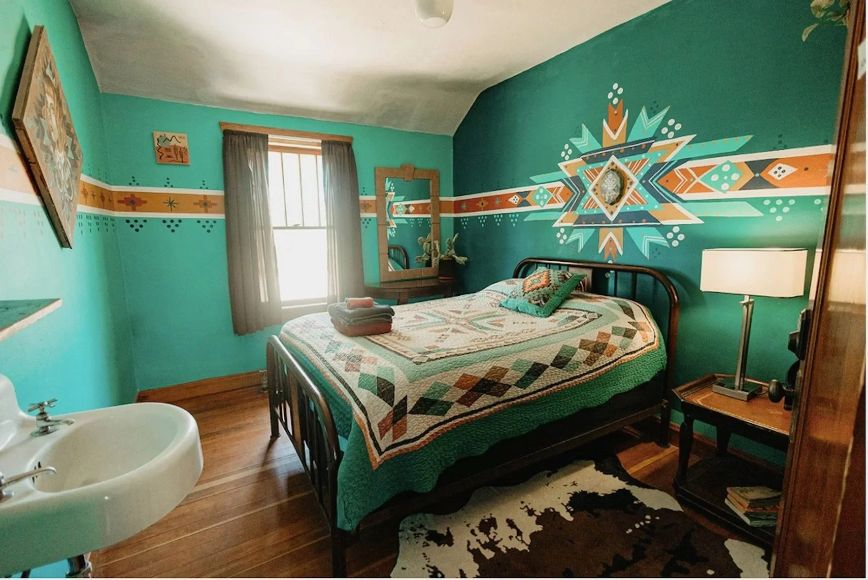 A bedroom with teal walls featuring southwestern style geometric mural art, a black metal bed with a quilt, a bedside table with a white lamp, a tall mirror with a wooden frame, a window with curtains, a cowhide rug on wooden floor, and a small sink in the corner.