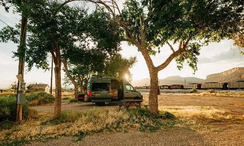 A small black van parked under trees in a rural setting, with a train and mountains visible in the background during sunset.