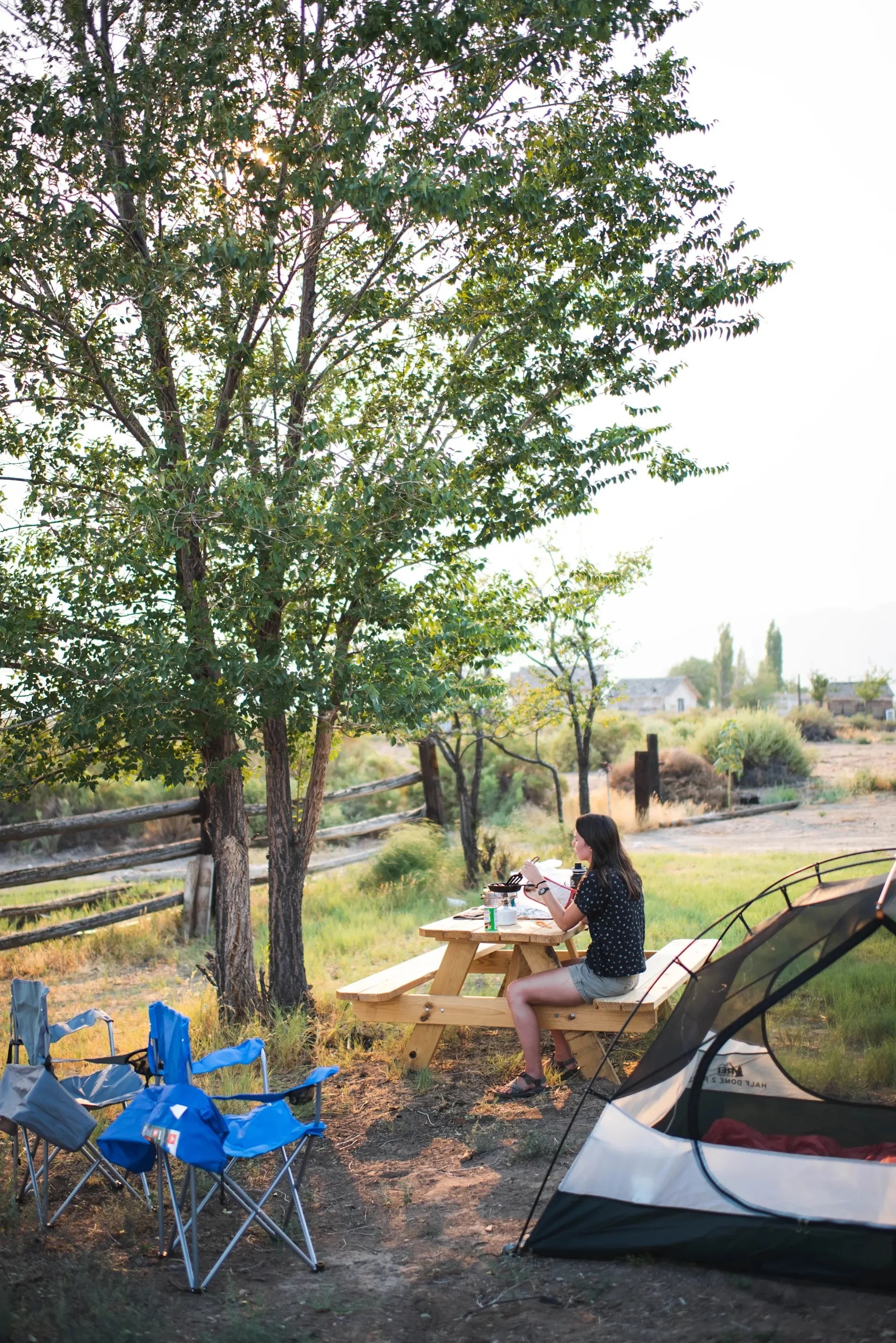 A woman sitting at a picnic table outdoors, enjoying a meal. Nearby are blue foldable chairs and a tent, with trees and houses in the background.