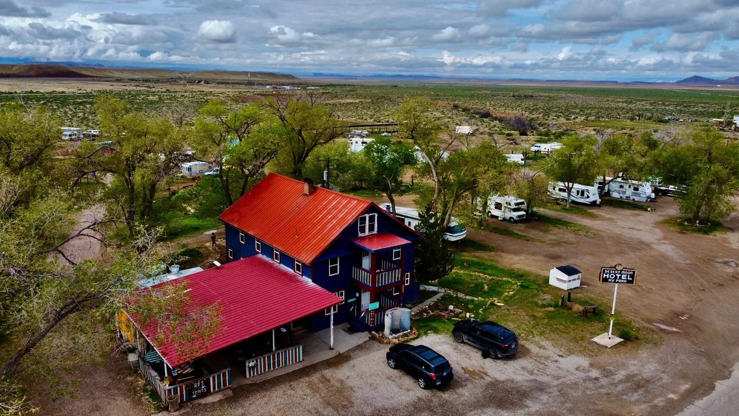 Aerial view of a rural campground with RVs, trees, and a blue building with a red metal roof; sign reads 'Desert Moon Hotel RV Park'.