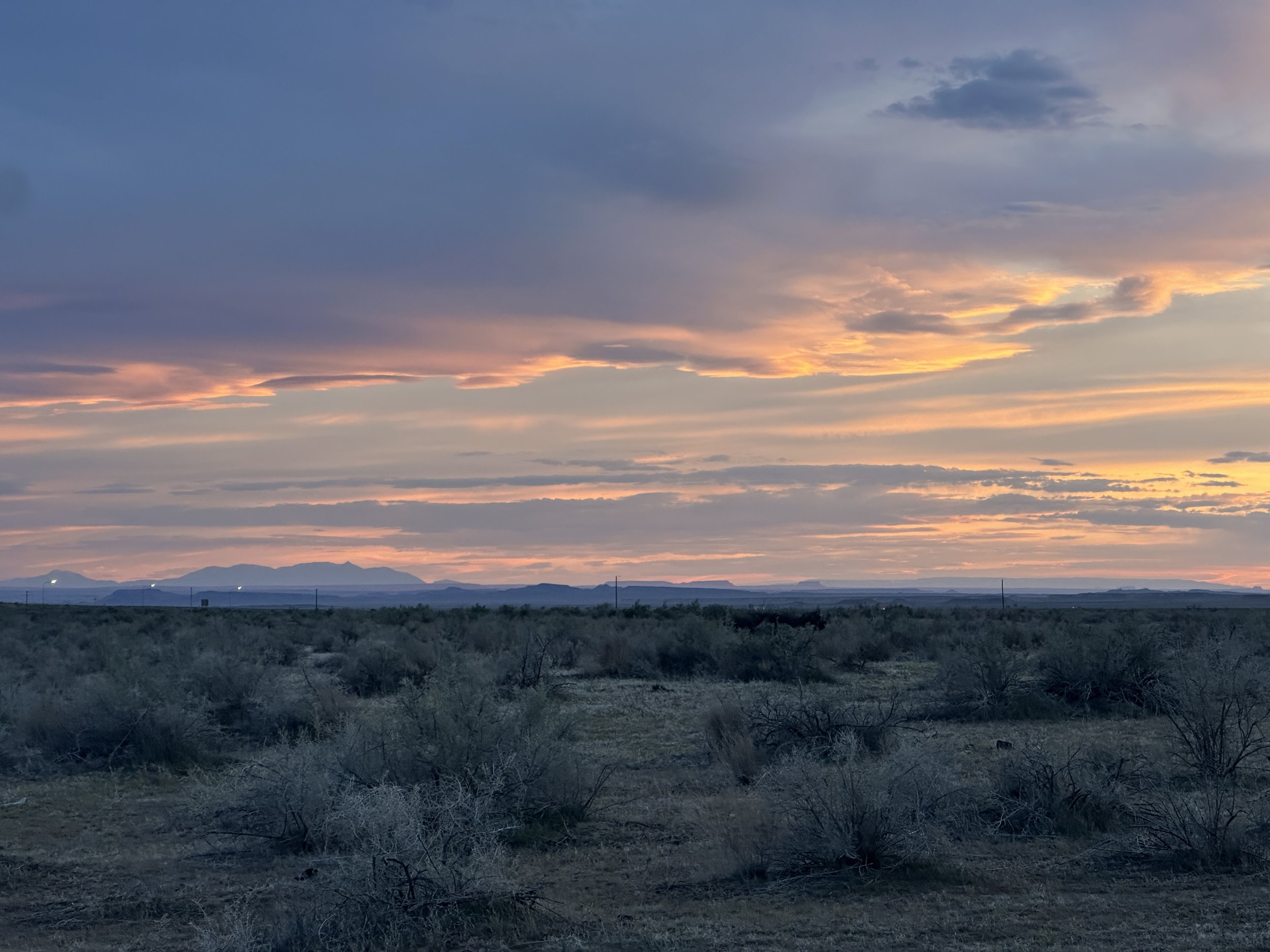 A vast desert landscape at sunset with a colorful sky and distant mountains.
