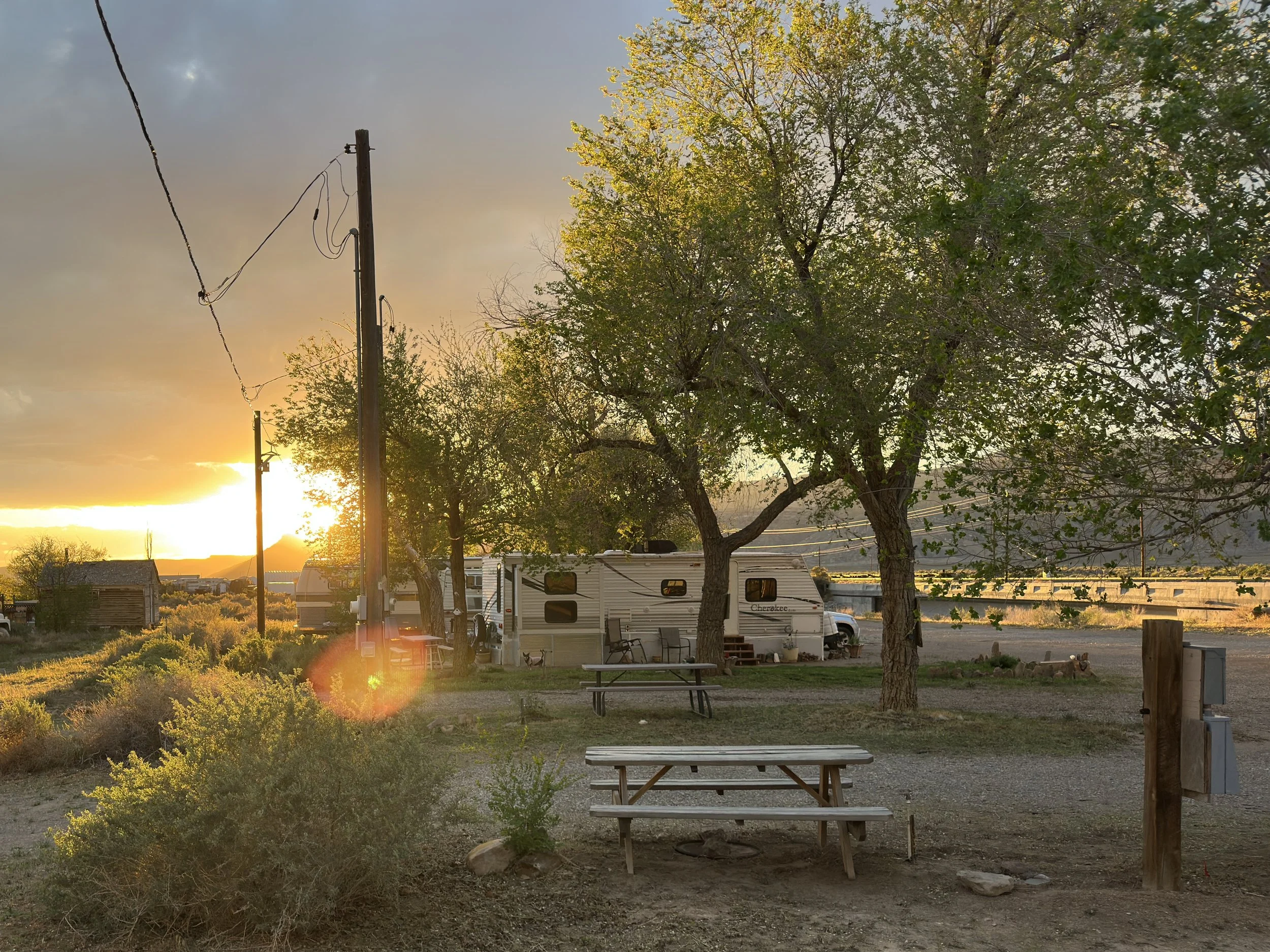 Sunset at a campground with trees, a picnic table, portable chairs, and an RV trailer. Power lines run through the scene, and the sky has a mix of clouds and clear areas, with the golden light of the setting sun.
