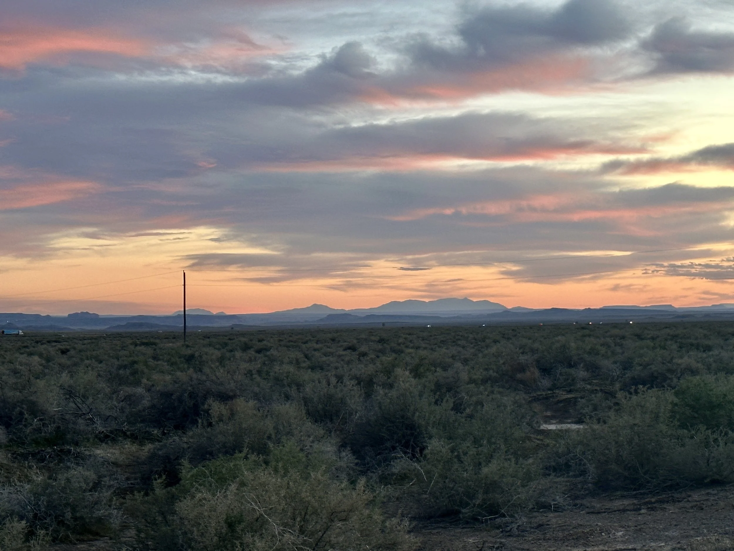 A desert landscape at sunset with a sky filled with clouds in shades of pink, purple, and yellow. Mountains are visible in the distance, with sparse vegetation and a single utility pole in the foreground.