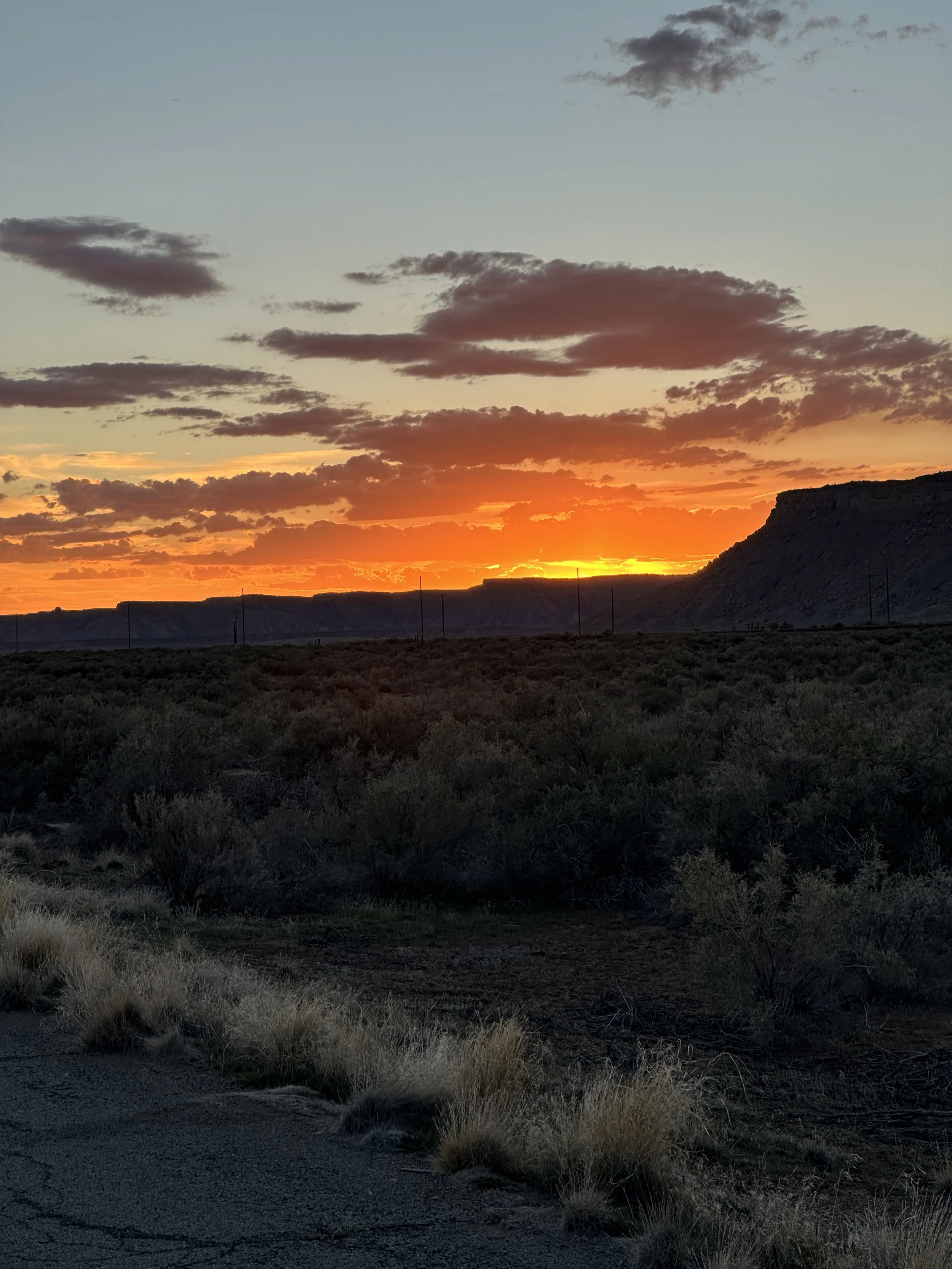 A sunset over a semi-arid landscape with low shrubs and a hill on the right side. The sky has scattered clouds illuminated with orange and yellow hues from the setting sun.
