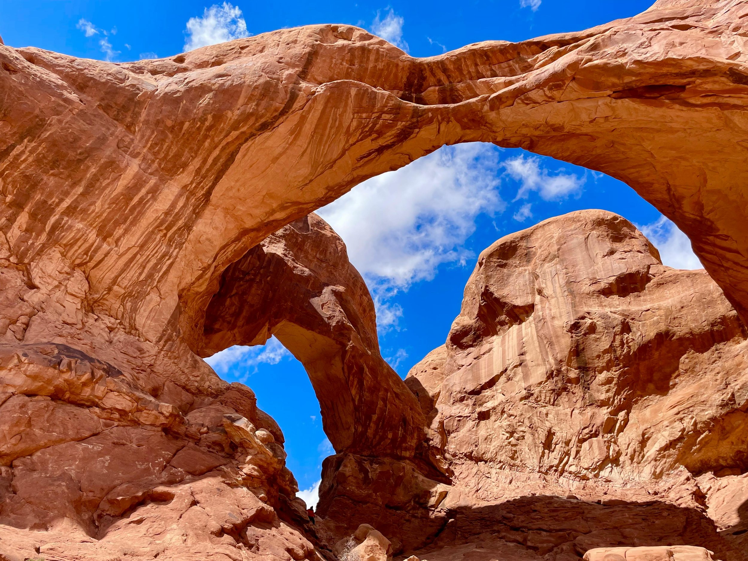 A natural rock formation with large red and orange sandstone arches and cliffs, under a blue sky with scattered clouds.