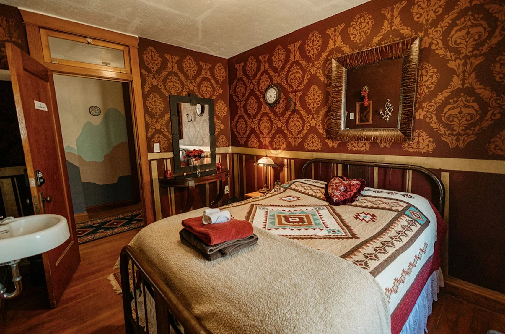 A cozy bedroom with vintage decor featuring a double bed with a quilted bedspread, a heart-shaped pillow, and neatly folded towels on a beige blanket. The walls are adorned with rich, red damask wallpaper and decorated with a large framed mirror, a small clock, and a wall sconce. There is a wooden door leading to a hallway, and a small sink is visible in the corner.