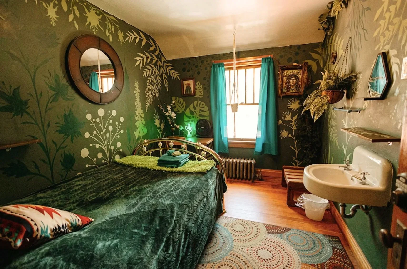 A bedroom with green leafy patterned wallpaper, a metal bed with green bedding, a small nightstand with a neon cactus light, a window with teal curtains, a corner sink, and wall art, with a multicolored circular rug on wooden floor.