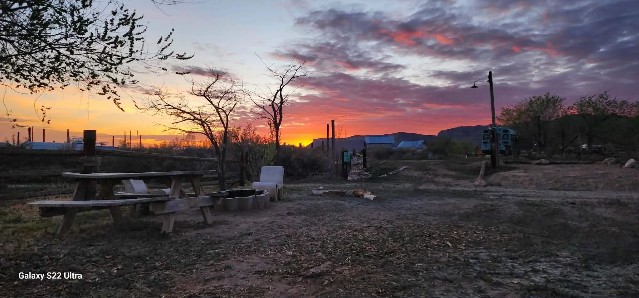 Sunset over a rural outdoor area with leafless trees, a picnic table, a chair, and a mountain in the distance, with a cloudy sky illuminated by orange and purple hues.