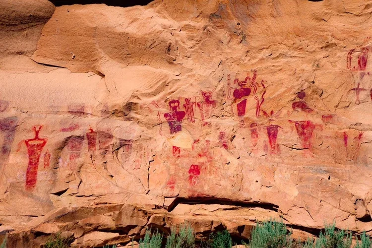 Ancient petroglyphs painted in red on a reddish-brown rock face depicting humanoid and animal figures.