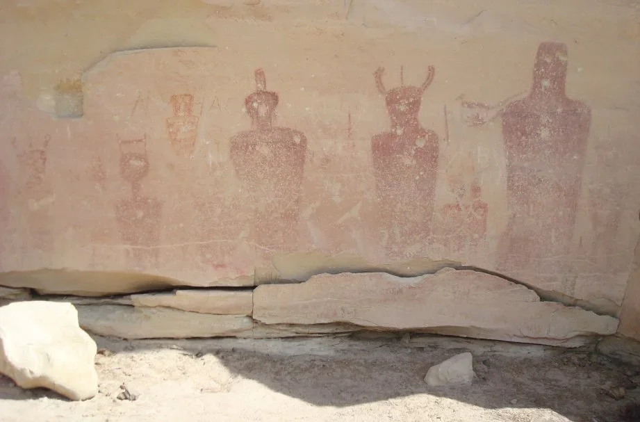 Ancient rock wall with faded red/pink petroglyphs of four humanoid figures, some with headdresses or horns, in a desert setting.