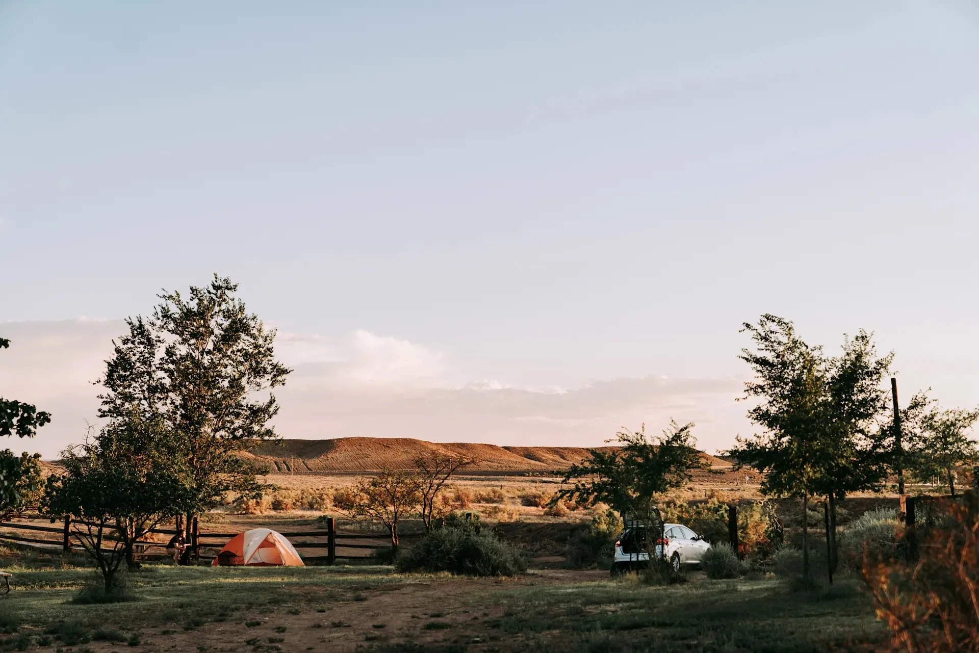 A scenic outdoor landscape during sunset with trees, a tent, a white car, and a rural fence in the foreground, and rolling hills under a clear sky in the background.