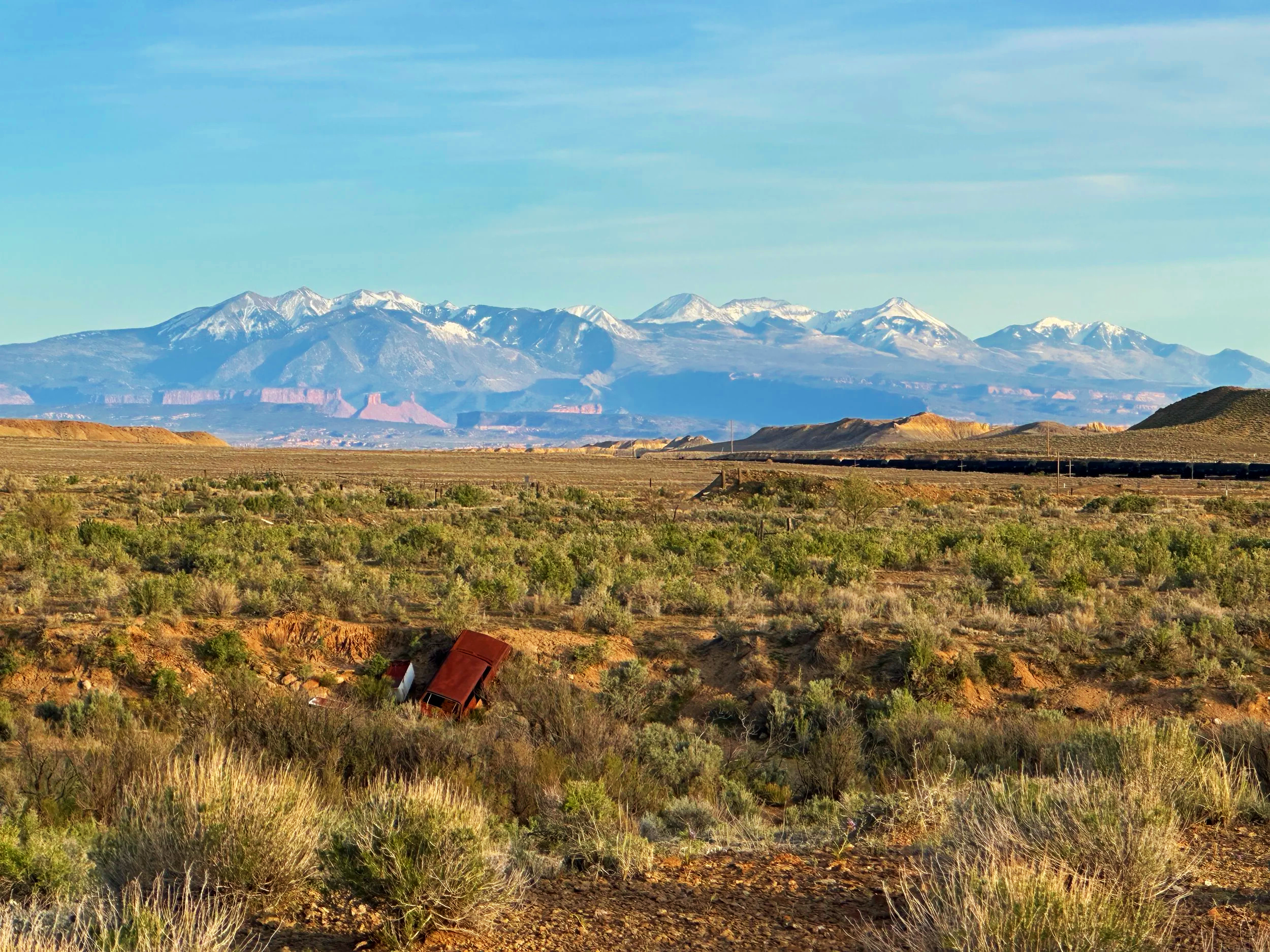 Desert landscape with distant snow-capped mountains, sparse vegetation, and abandoned vehicles in the foreground.
