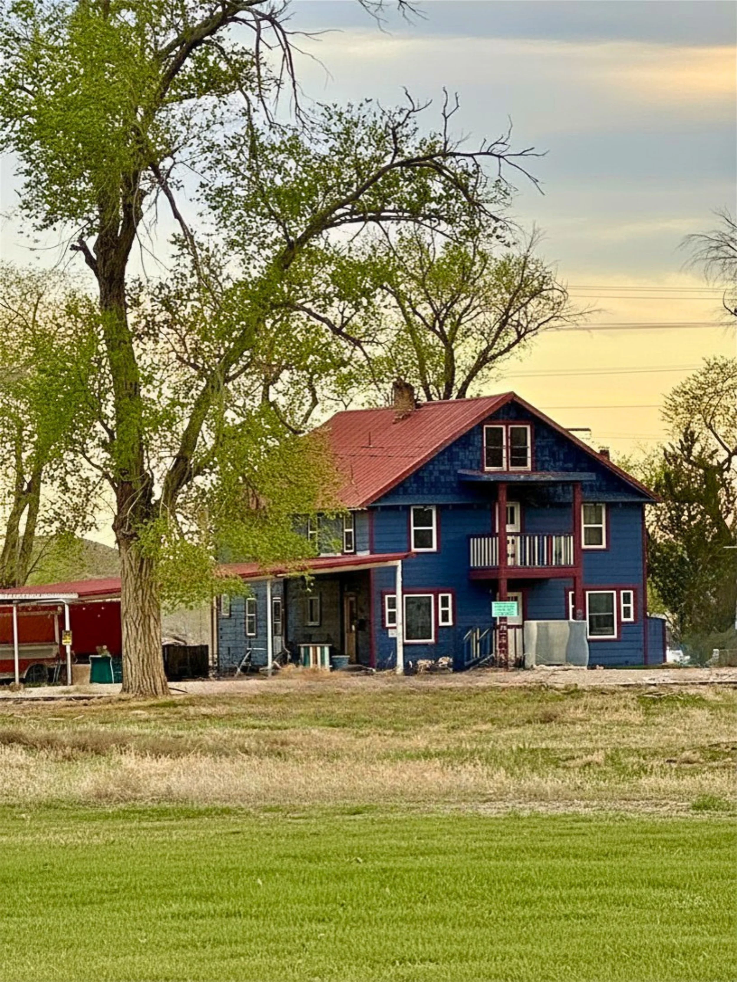A blue house with a red roof, multiple windows, trees surrounding it, and a grassy lawn in the foreground.