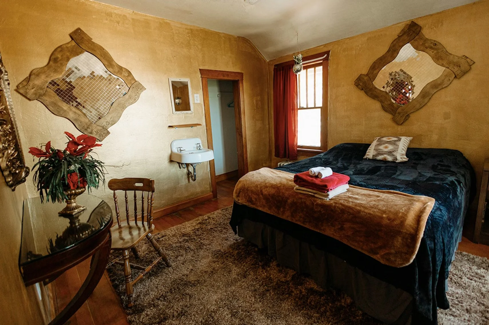 A cozy bedroom with yellow textured walls, a large bed with a dark blue cover, and a brown blanket at the foot. On the bed, there are neatly folded towels. Two asymmetrical decorative mirrors hang on the wall, and a small window with reddish curtains lets in natural light. A vintage-style sink is mounted on the wall near the door, and a small wooden table with two chairs and a potted plant with red flowers are in the room.