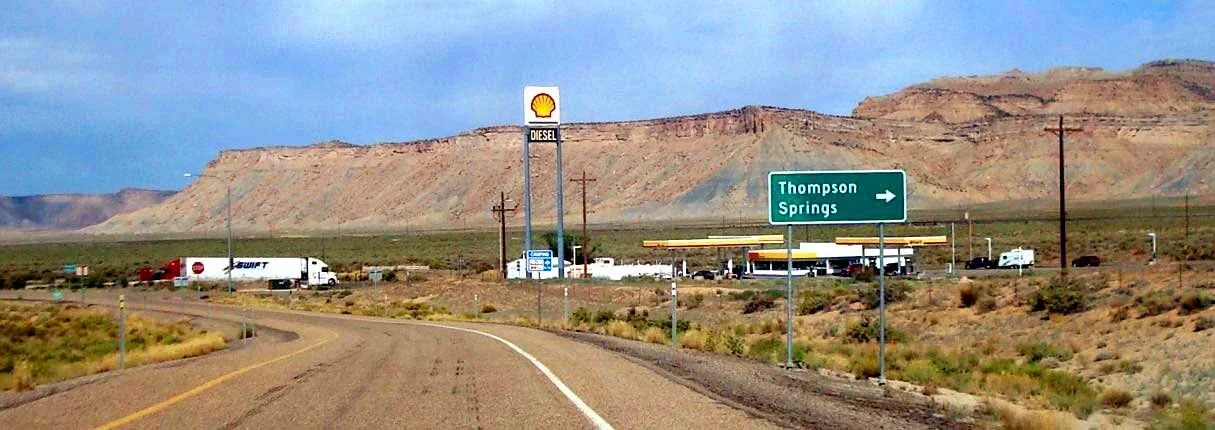 Desert highway scene with a Shell fuel station, a sign pointing to Thompson Springs, and a truck on the road with rocky hills in the background.