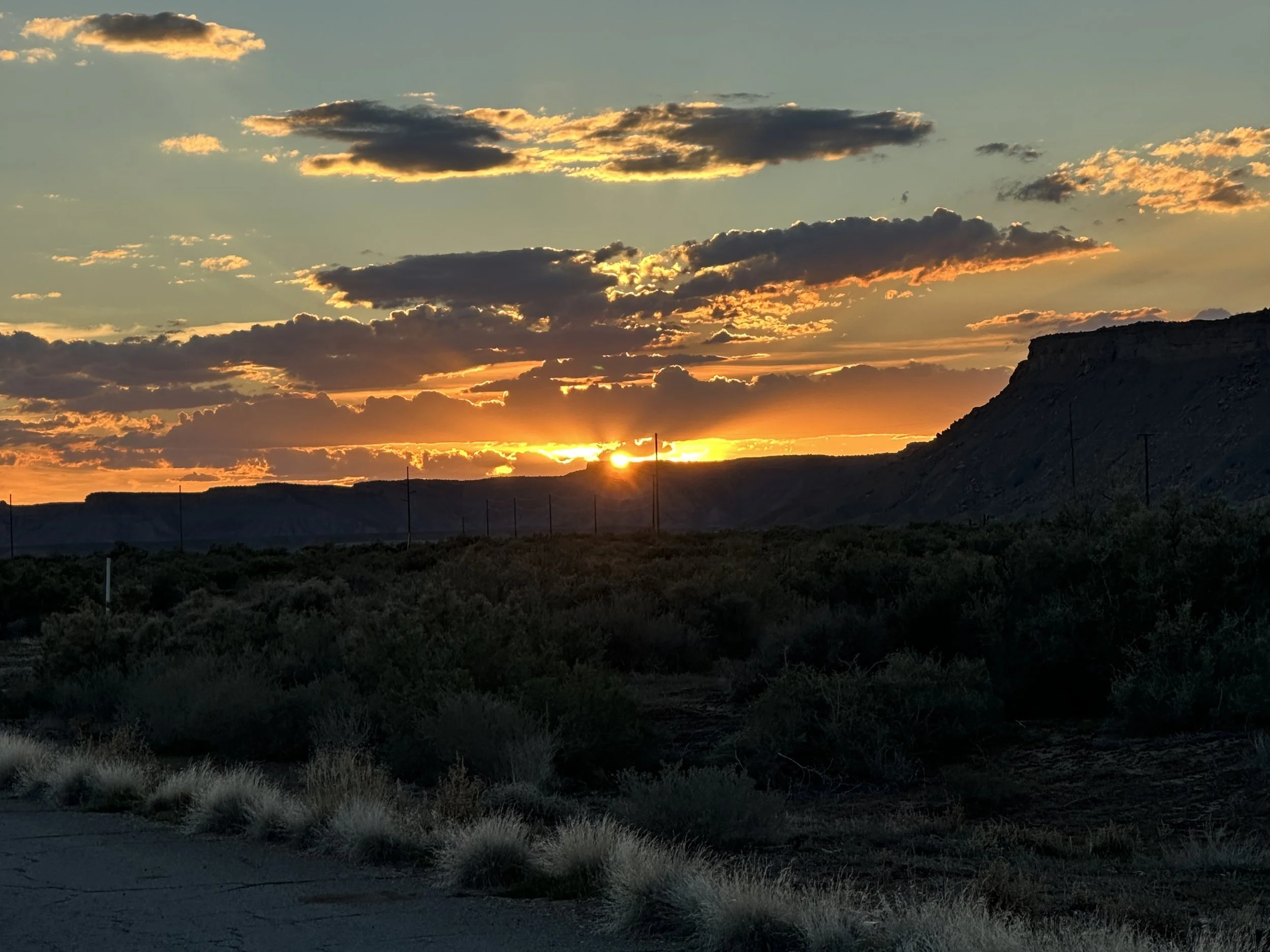 Sunset over a landscape with hills and sparse vegetation, with clouds in the sky illuminated by the setting sun.