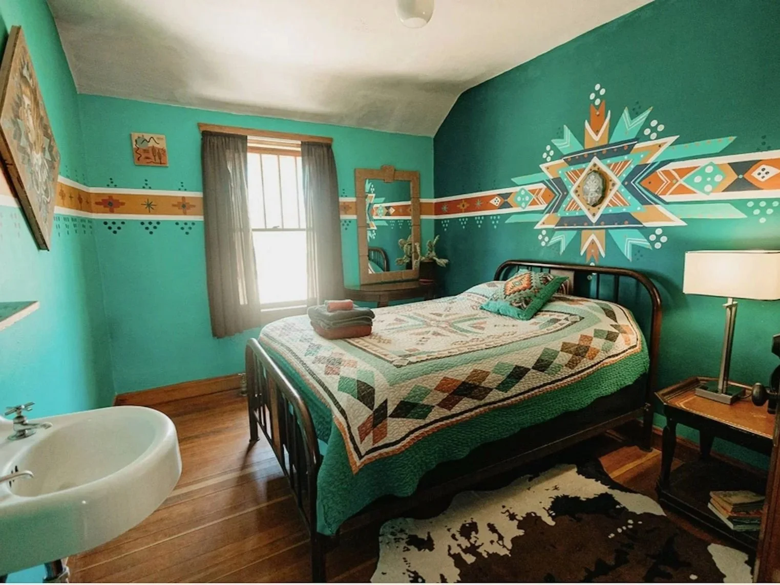 A bedroom with turquoise walls featuring a large geometric mural behind the bed, a wooden floor, and a window with brown curtains. The bed has a colorful quilt and pillows, and there is a nightstand with a lamp and a mirror above it. Some towels are folded on the bed and a cowhide rug is on the floor.