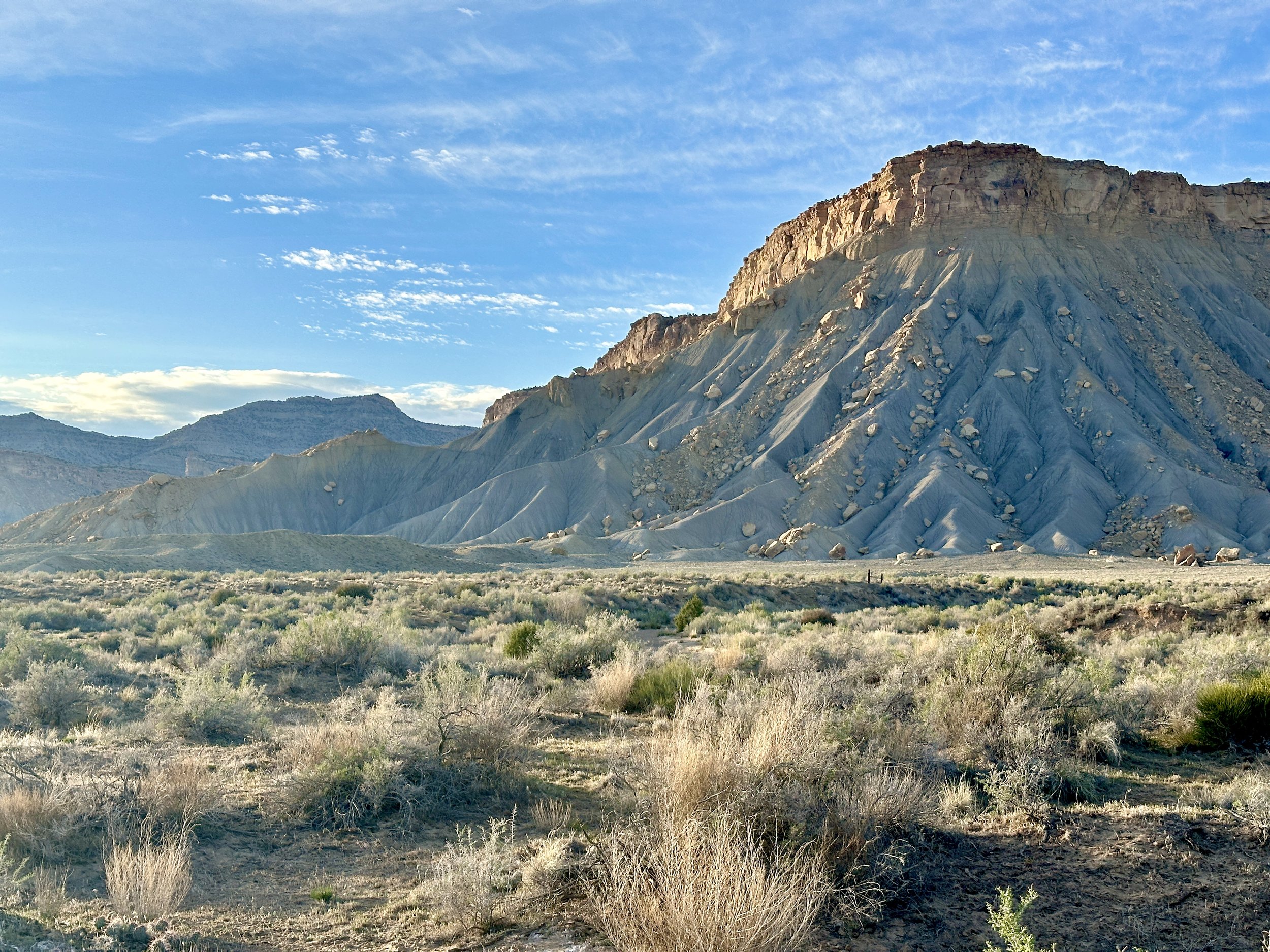 A rugged, barren mountain landscape with a large, flat-topped mountain in the center under a partly cloudy blue sky. The foreground consists of dry grass and sparse shrubbery.