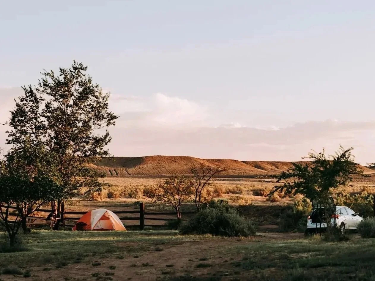 Camping scene with an orange and white tent and a white car parked on a dirt area near trees and bushes, with open land and hills in the background during sunset.