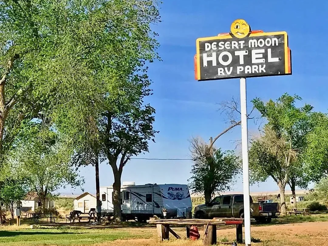 A sign for Desert Moon Hotel RV Park with a smiling face emoji on top, set against a blue sky and surrounded by trees with RVs and vehicles in the background.