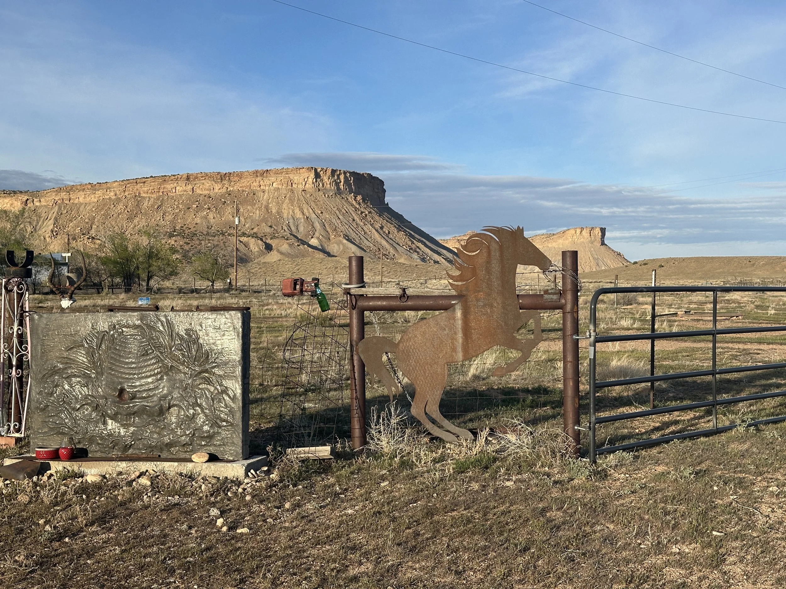 A desert landscape with a flat-topped mesa, some shrubs, and a metal sculpture of a horse's head in the foreground. There are some fences and metal objects near the sculpture.