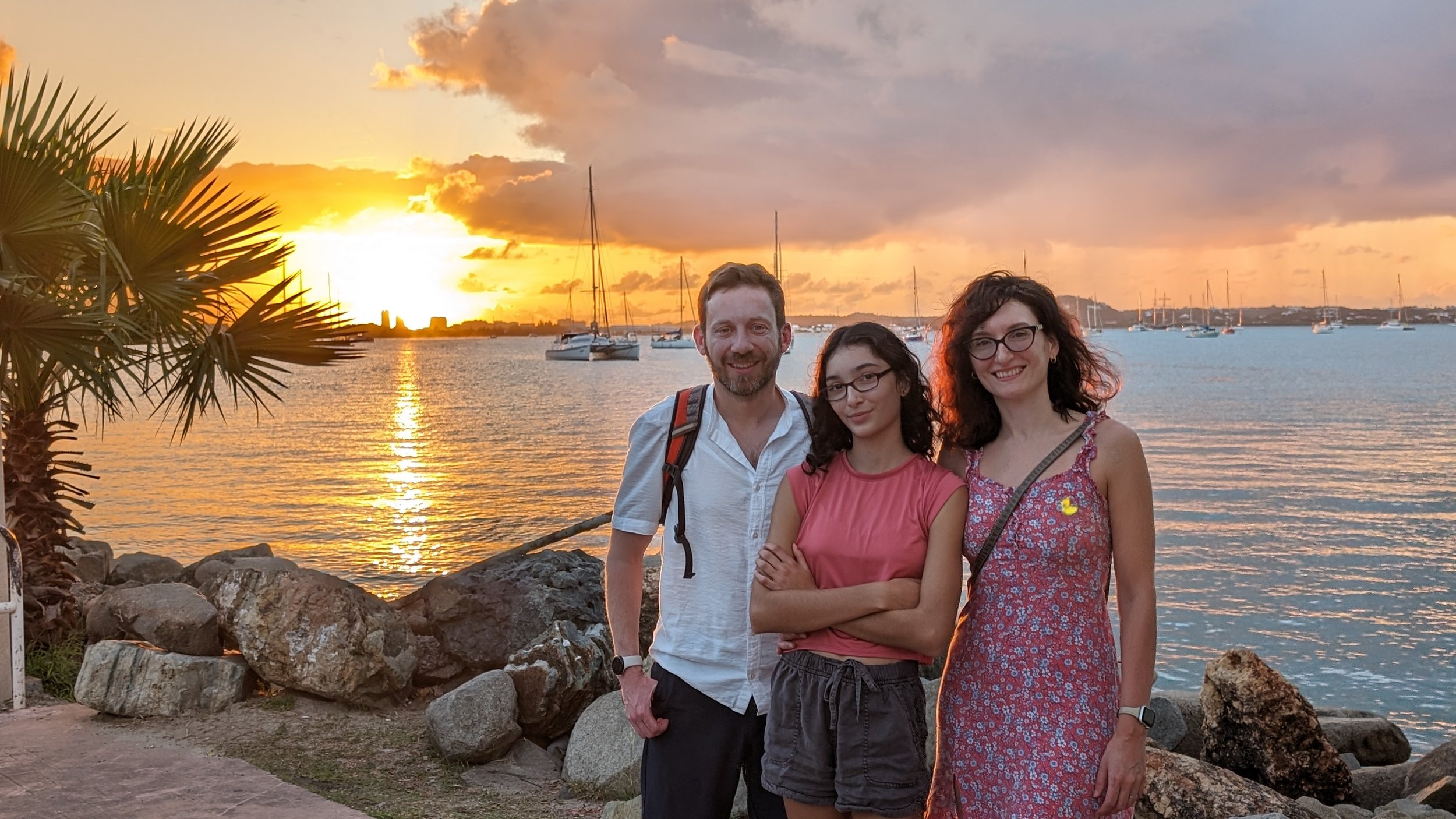 A family of three standing on a rocky shoreline at sunset, with boats in the water and clouds in the sky.