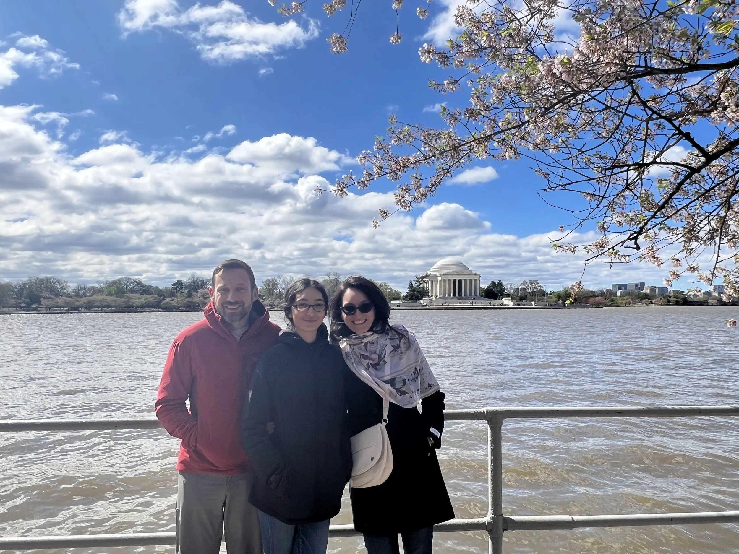 Three people standing in front of the Lincoln Memorial and reflecting pool in Washington D.C., with cherry blossom branches overhead and a partly cloudy sky.