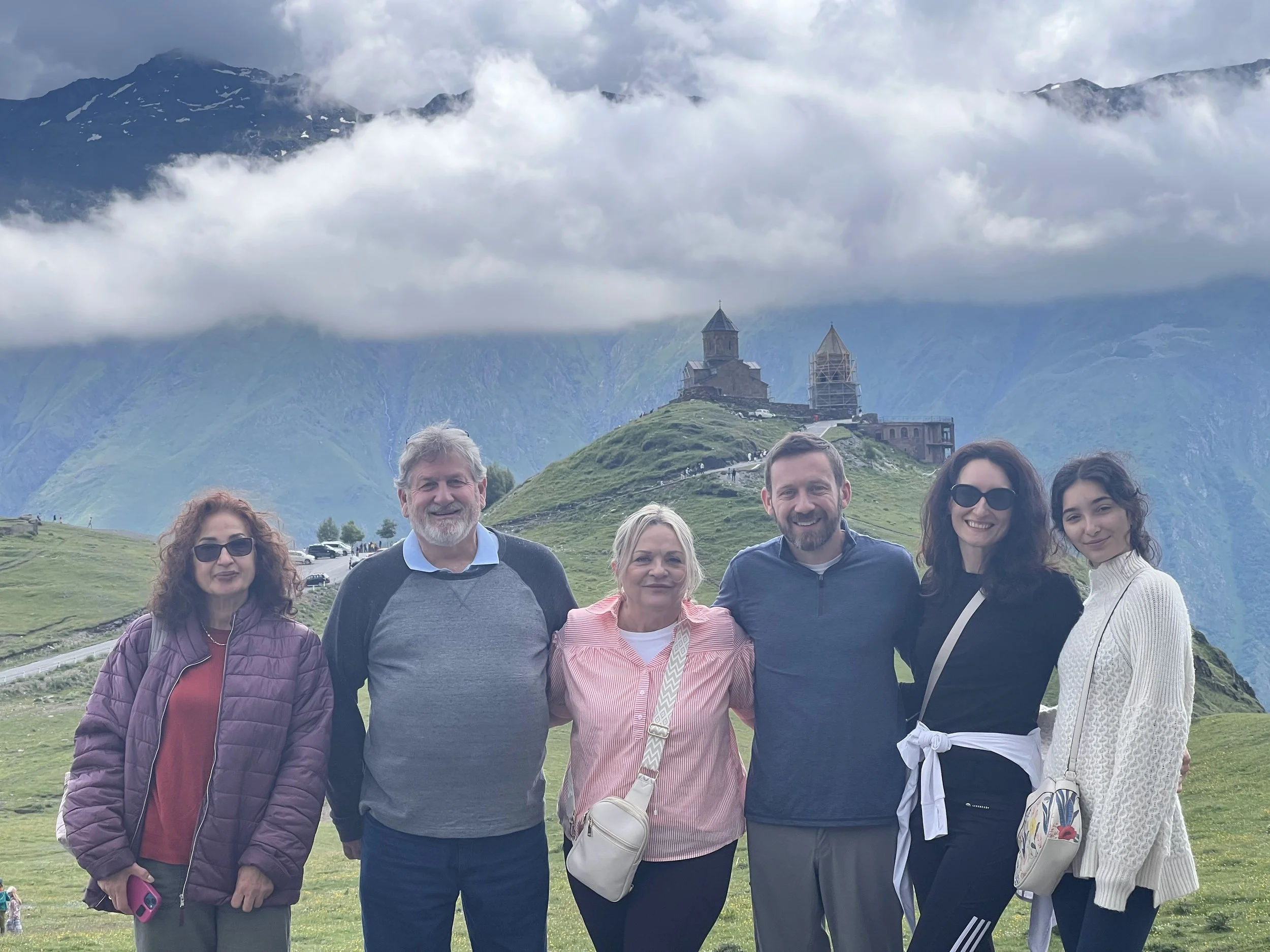 Group of six people standing together outdoors with mountains and ancient church buildings in the background, on a cloudy day