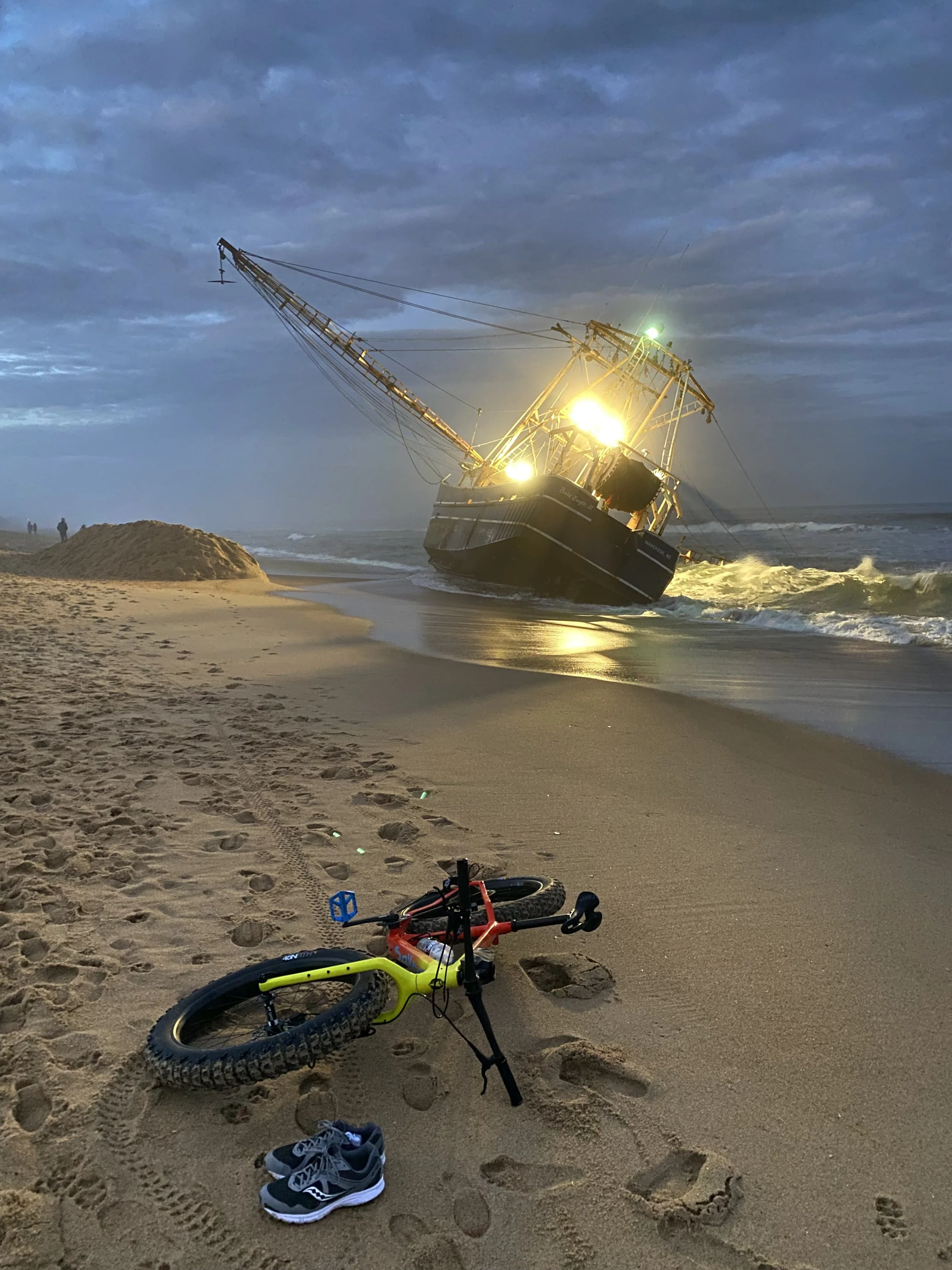 A fat tire bicycle lying on a sandy beach with a pair of shoes nearby. A shipwreck is visible in the background near the shoreline, with the ship tilted at an angle and illuminated by bright lights during dusk.