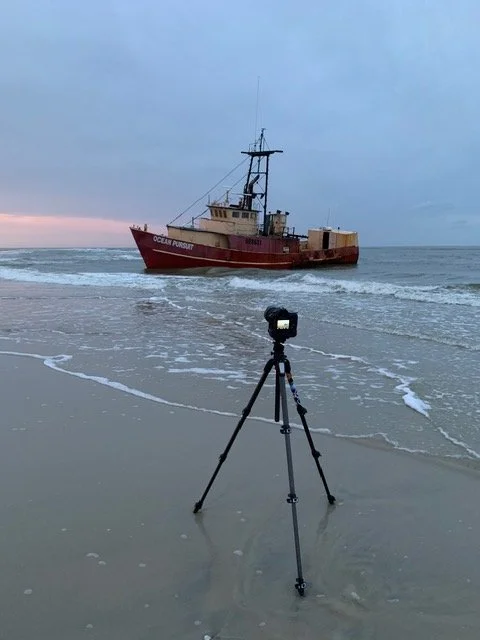 A red and white ship named Ocean Pursuit approaching the beach with a camera on a tripod set up on the shoreline in the foreground, during a cloudy day.