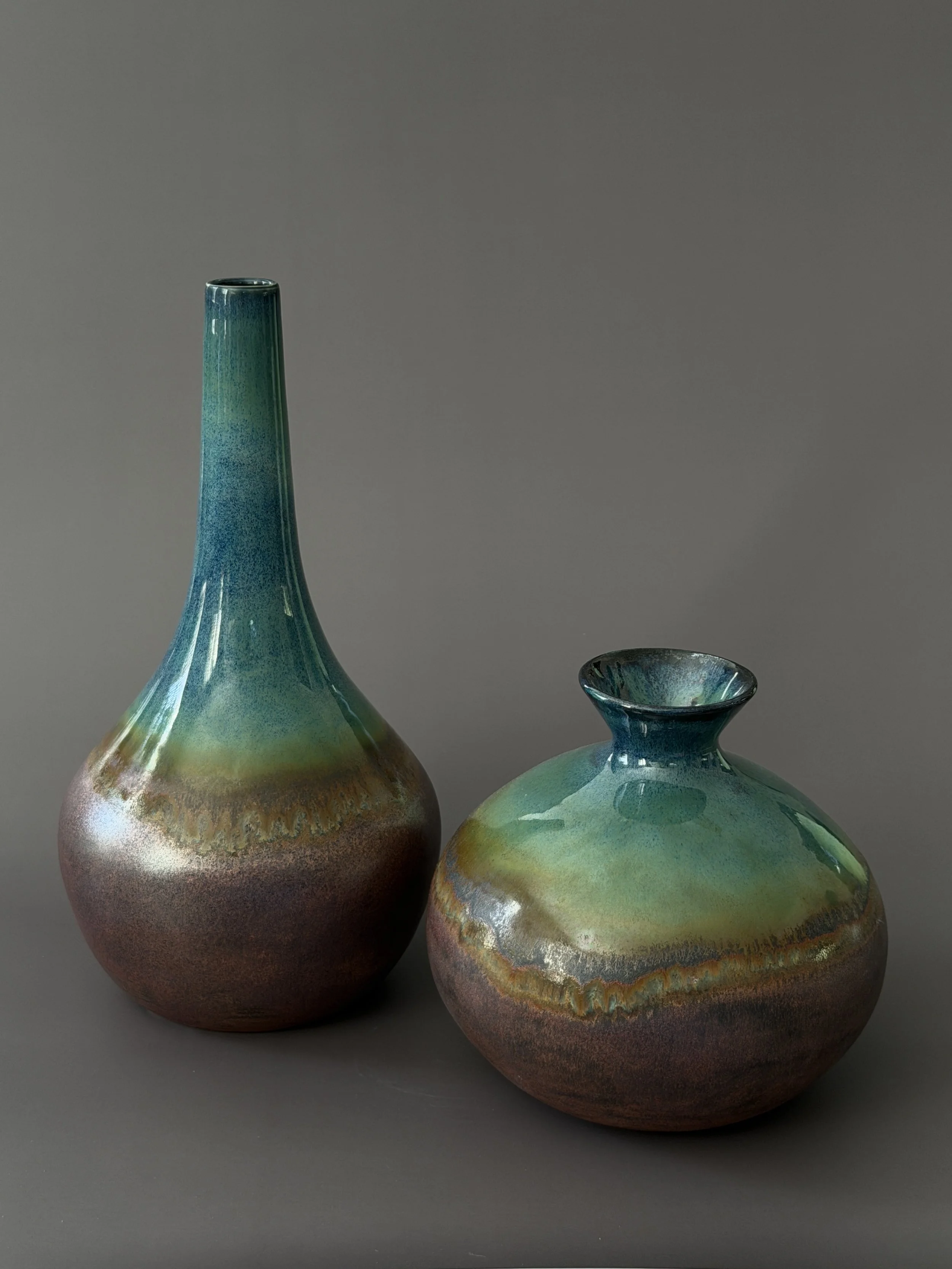 Two glazed ceramic vases with a gradient of blue, green, and brown colors, positioned against a plain gray background.