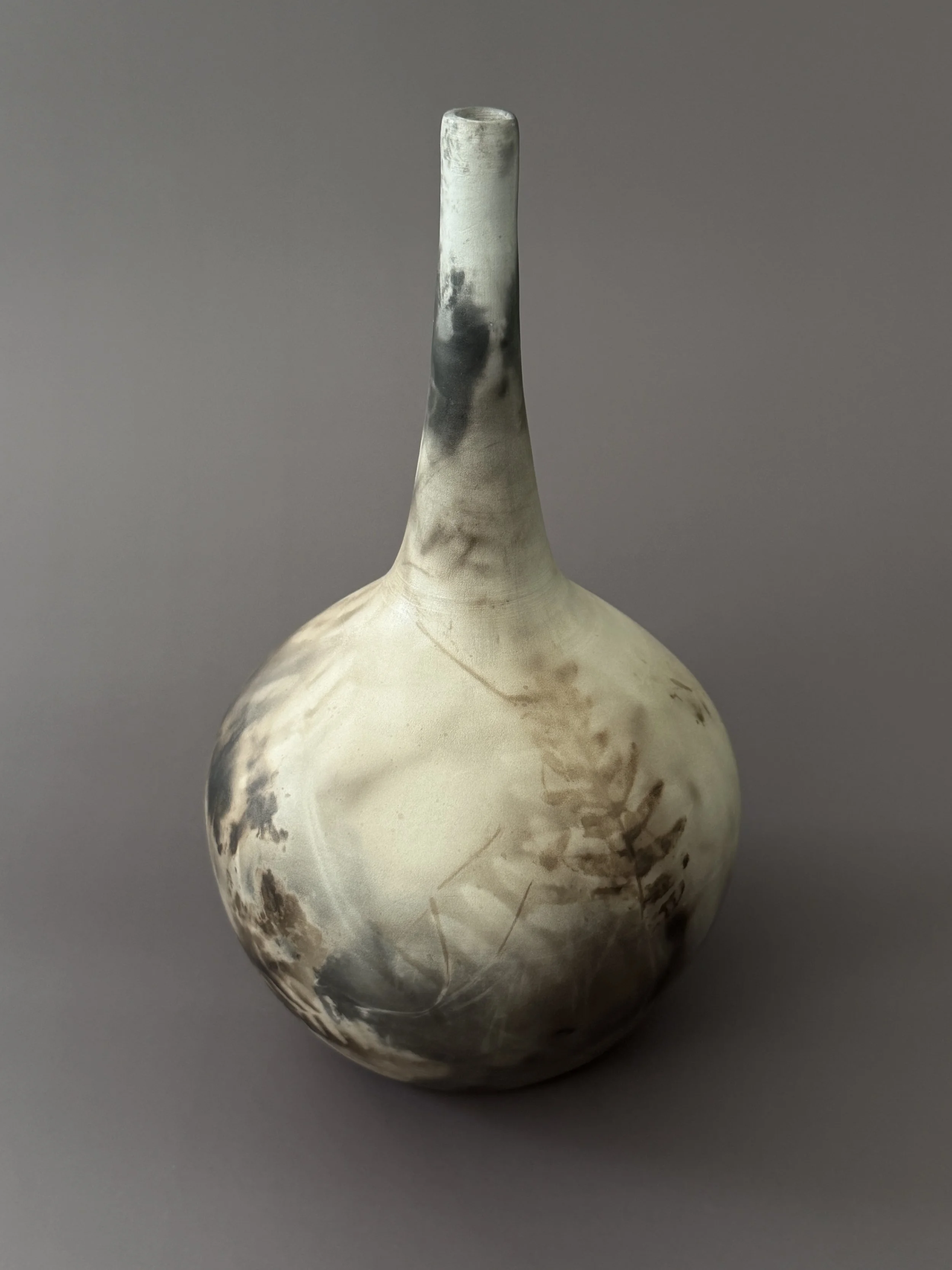 A ceramic vase with a long neck and a rounded body, featuring a black and white pattern made by leaves and smoke, set against a gray background.